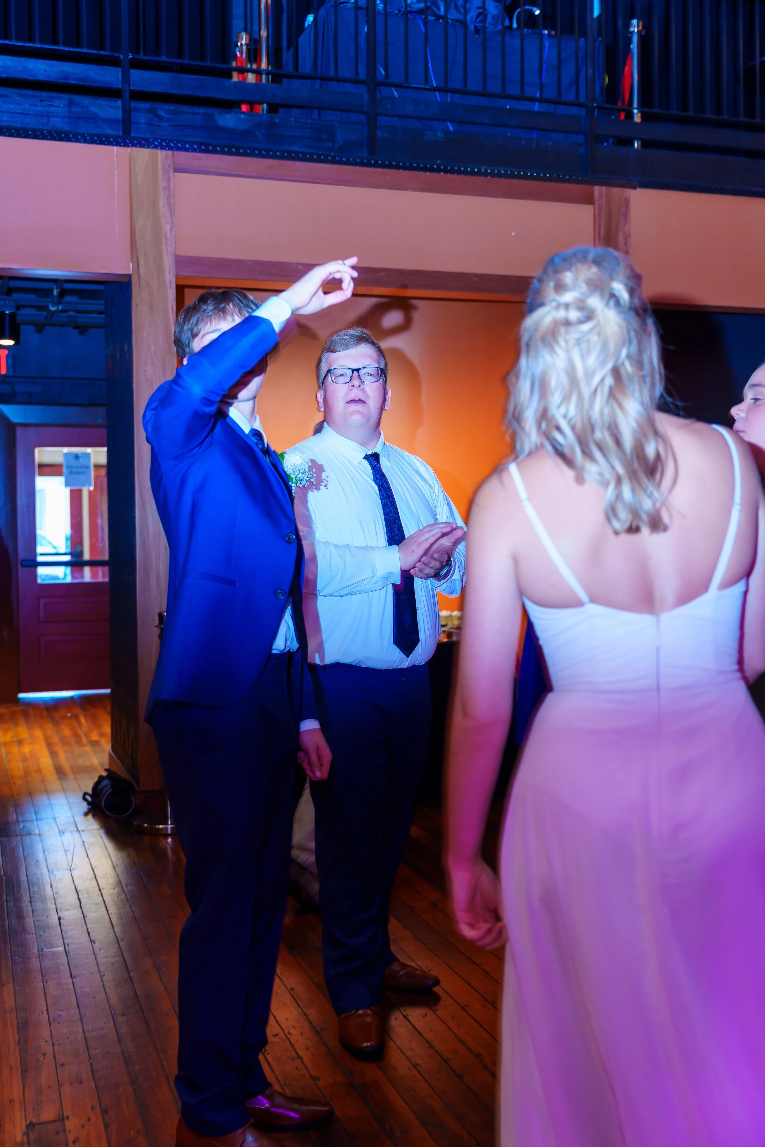 A group of people at a social gathering, with one man in a blue suit gesturing with his hand while others listen. The setting appears to be indoors with wooden flooring and warm lighting.