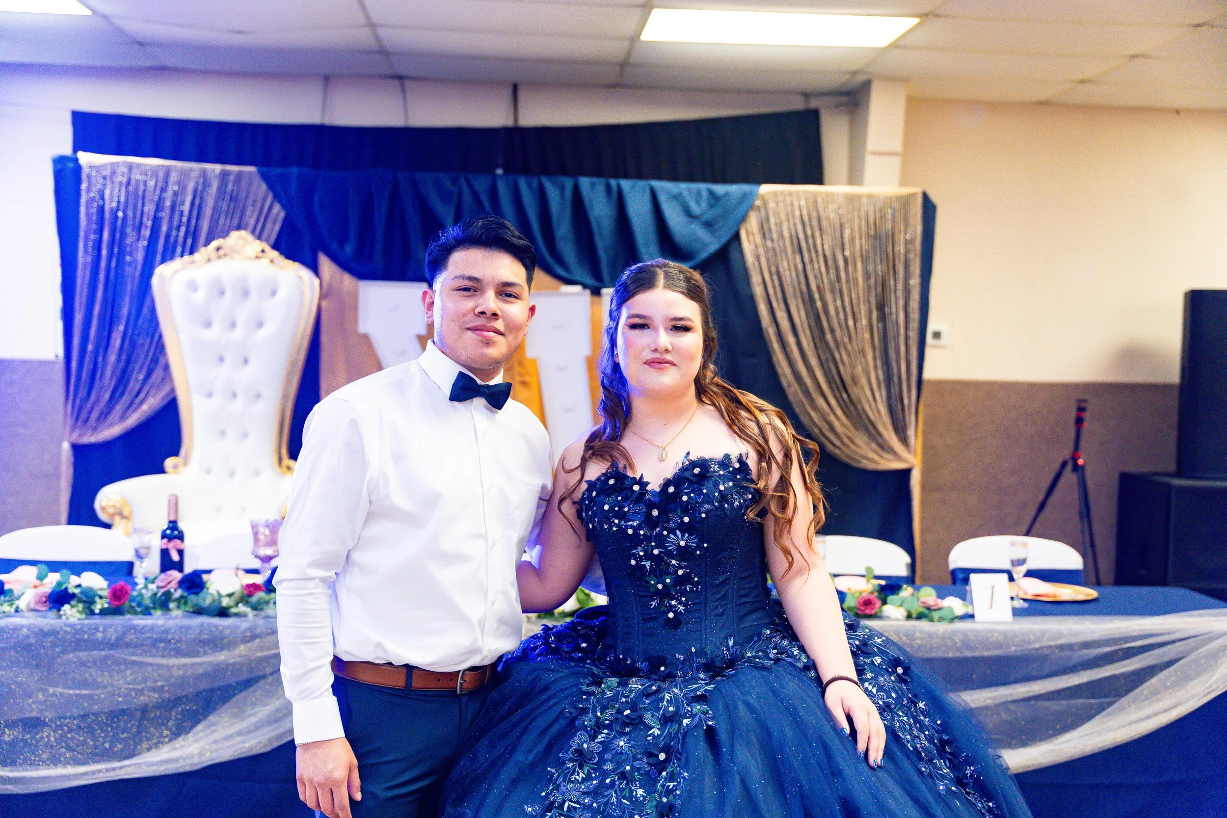 A young man and woman are posing together at a formal event, with the woman wearing a dark blue, embellished gown and the man in a white dress shirt with a black bow tie. They are standing in front of a decorated background with a dark blue drape and