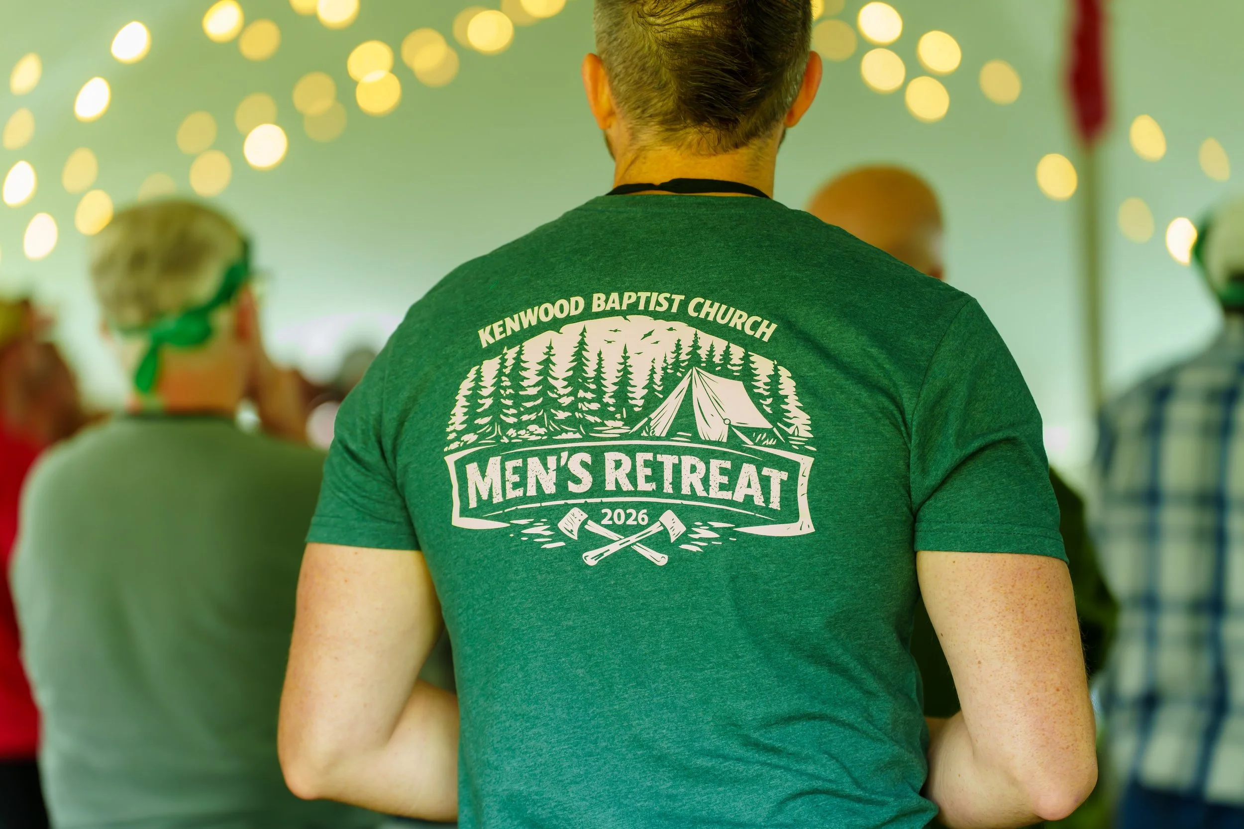 Back view of a person wearing a green T-shirt with the text 'Kenwood Baptist Church Men's Retreat 2026' and graphics of trees and crossed axes, indoors with blurred people and string lights in the background.