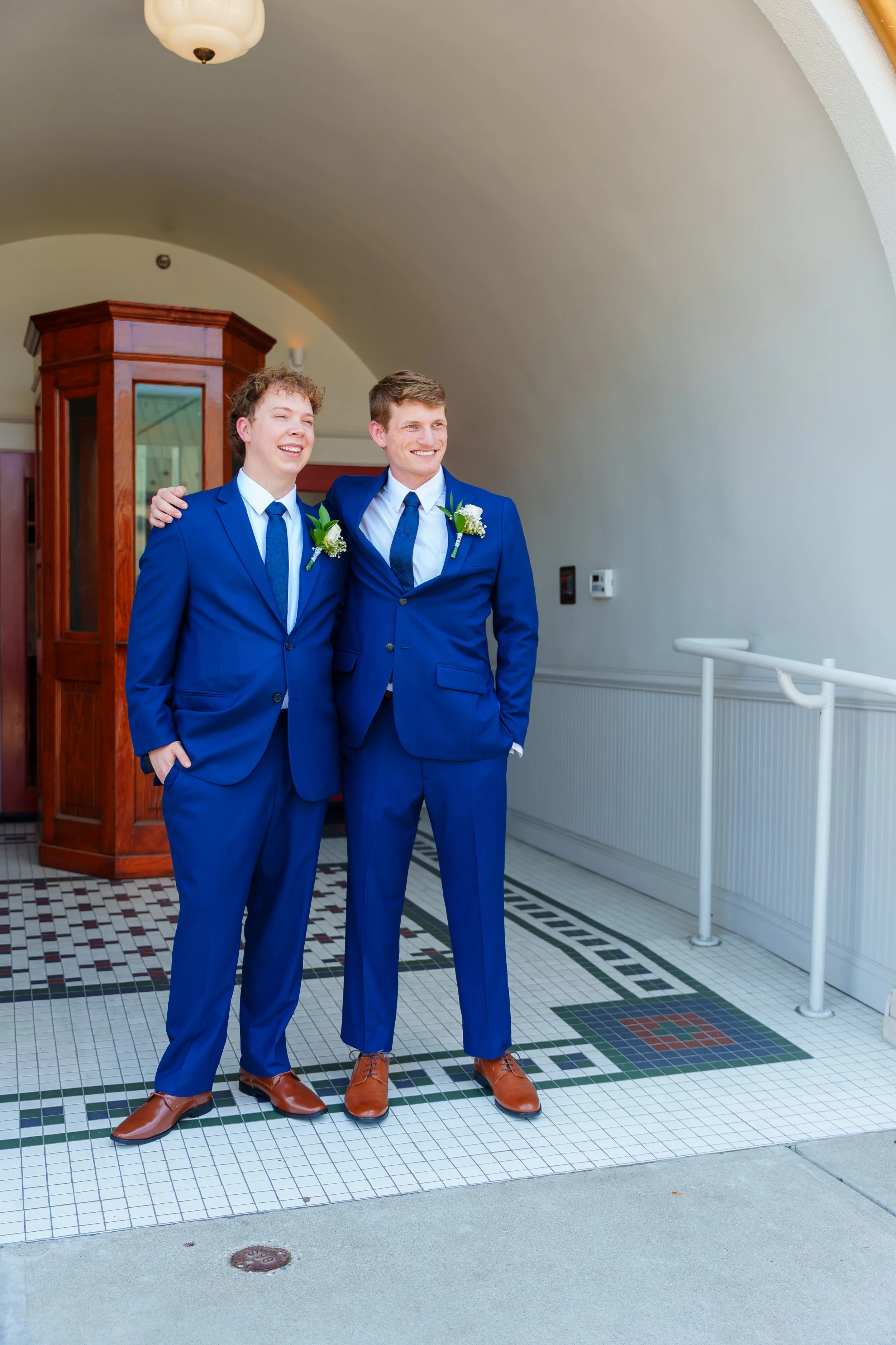 Two men dressed in blue suits and brown shoes standing in a tiled room, smiling and smiling, with one arm around each other, wearing boutonnières.