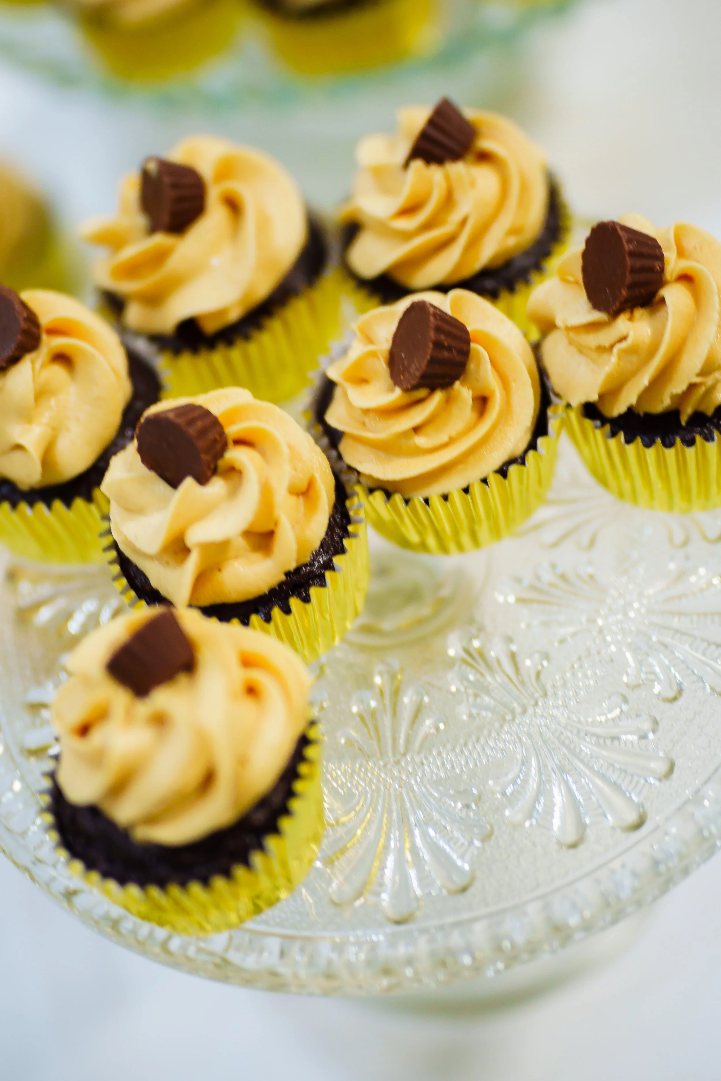 Close-up of yellow cupcakes with swirl frosting and small chocolate pieces on top, displayed on a decorative glass cake stand.