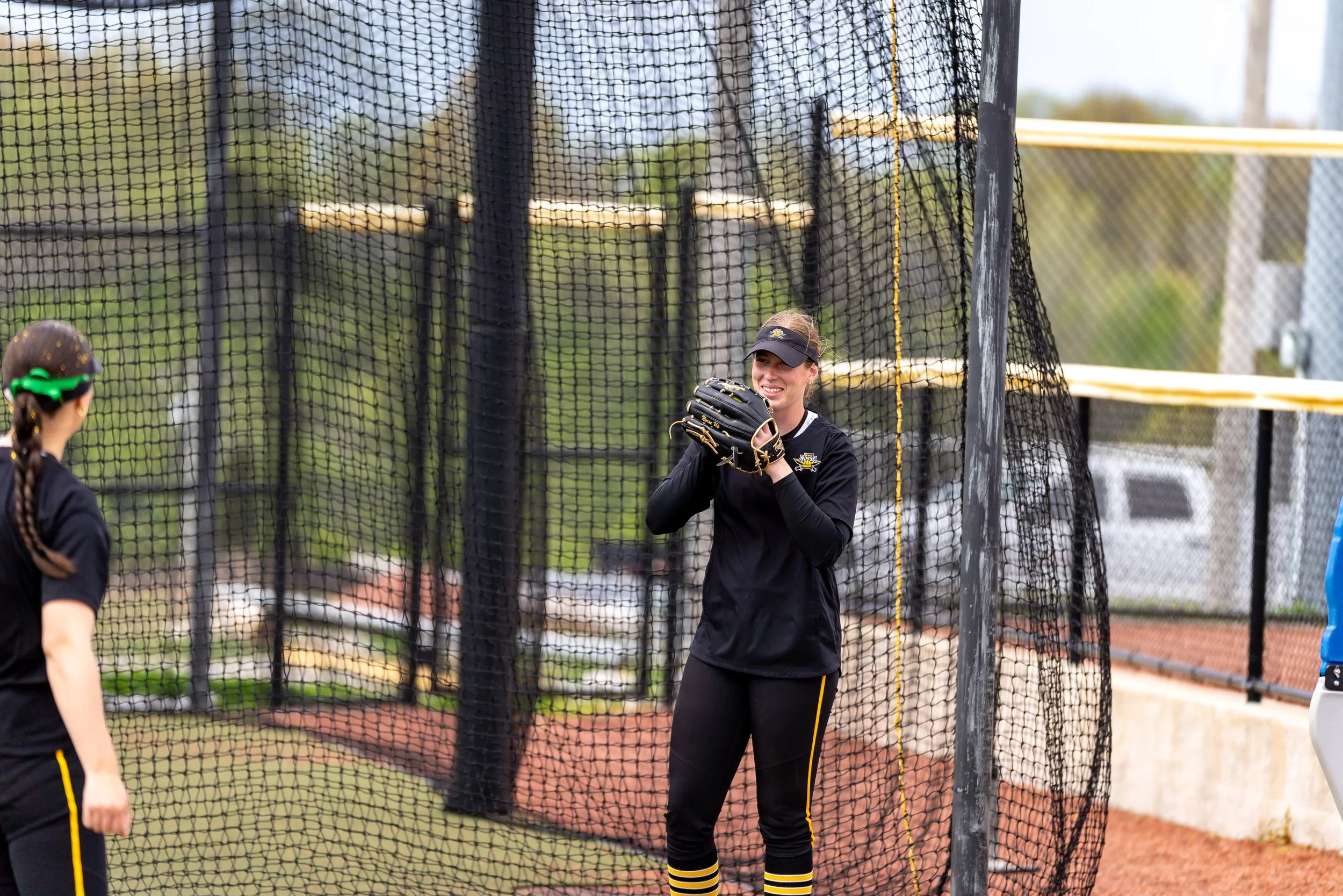 A female softball player standing near a batting cage, holding a glove and smiling, while another girl with a ponytail and sunglasses is seen from behind.