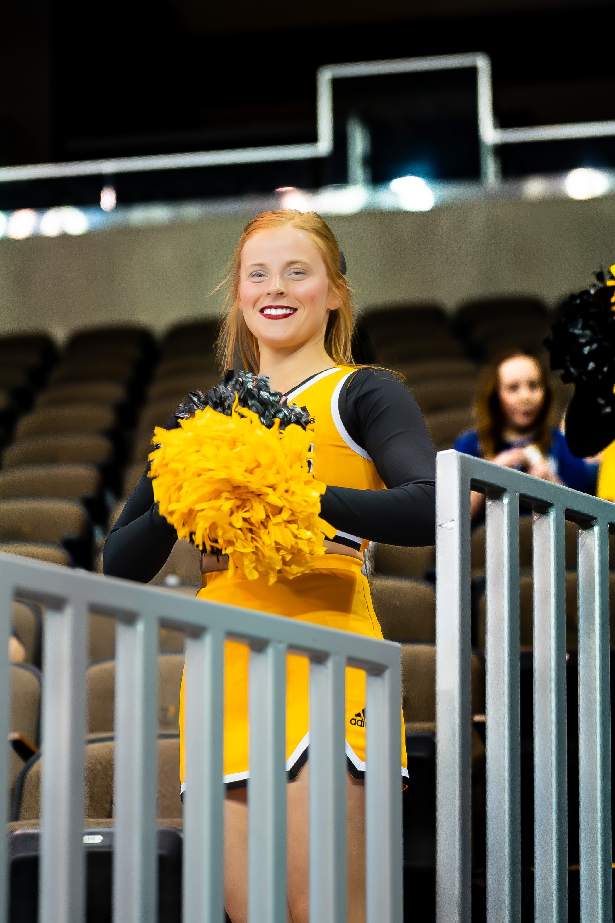Cheerleader in yellow and black uniform holding yellow pom-poms at an indoor sports event.