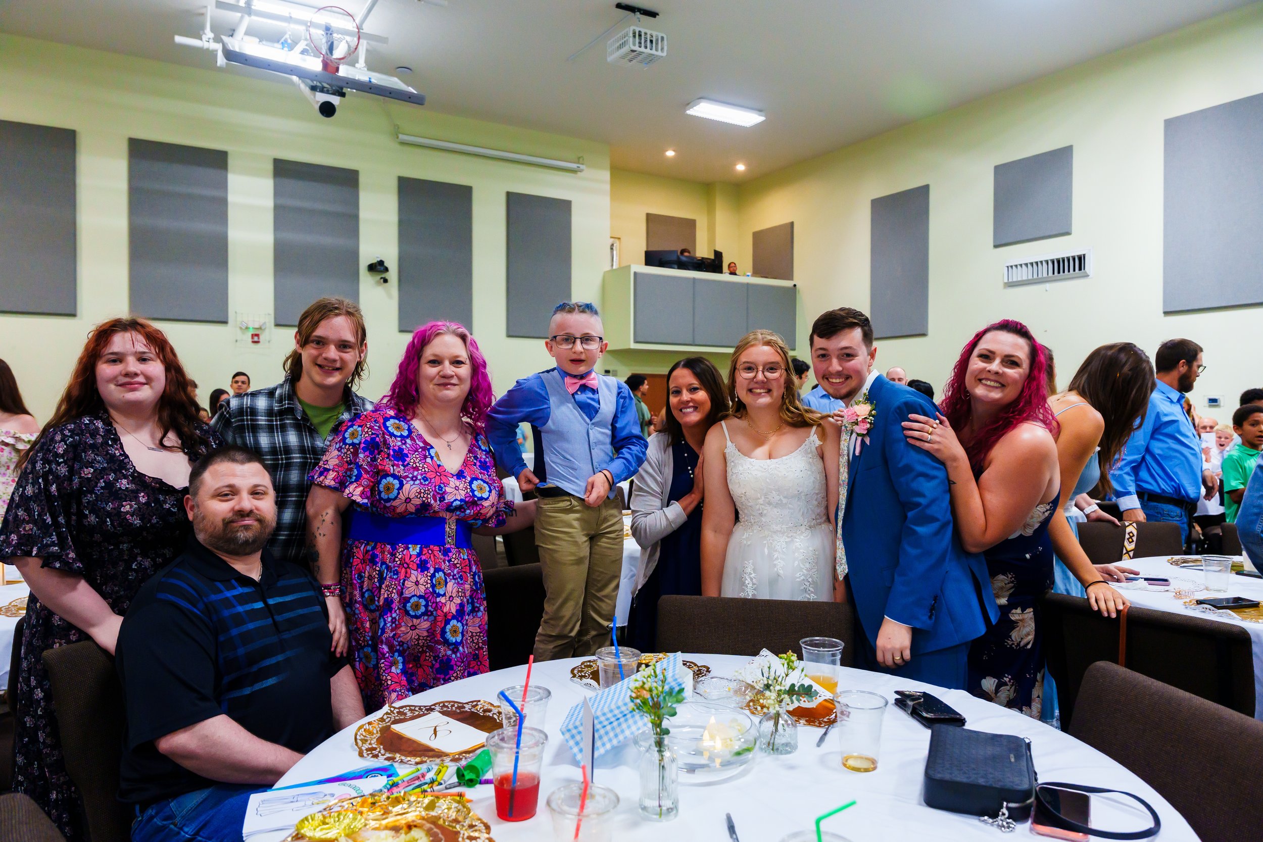 A group of people at a celebration, including men, women, and children. Some are dressed formally, with one woman in a white wedding dress and a man in a blue suit and boutonniere. They are standing close together smiling. The scene is in a banquet h
