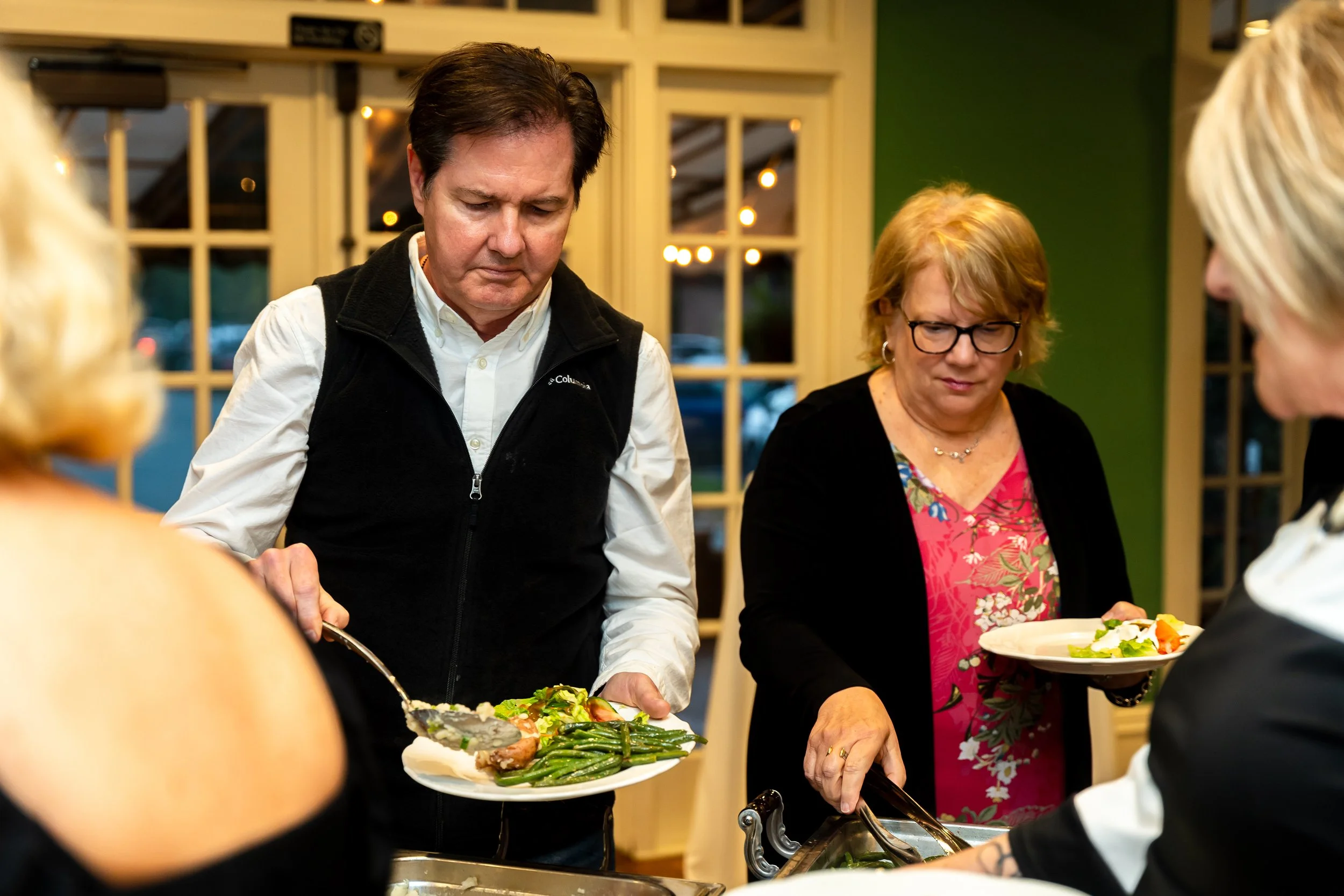 People serving themselves food at a buffet in a restaurant, with a man in a white shirt and vest and a woman in glasses and a floral top in the center.