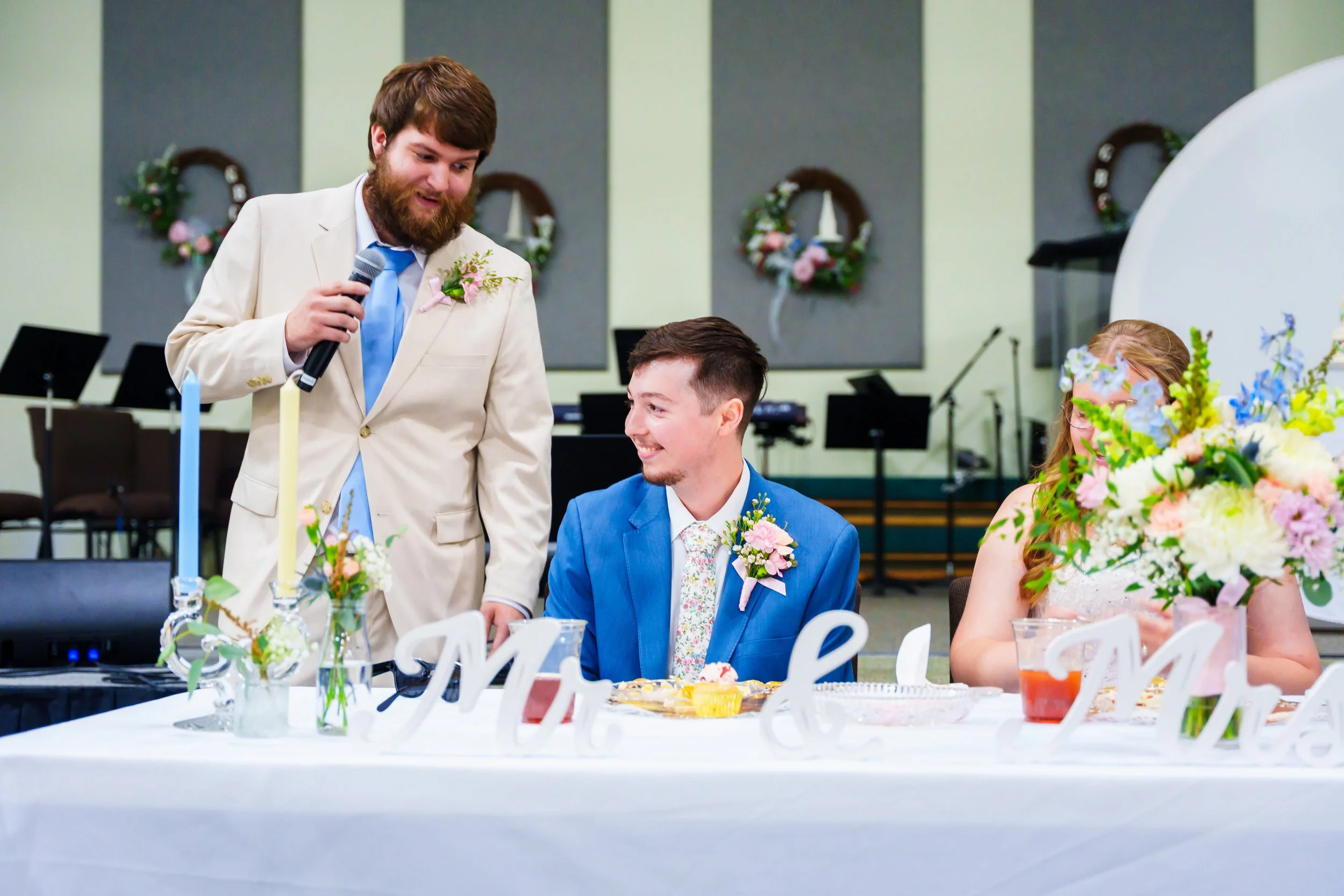 A wedding reception with a man in a cream suit and a blue tie speaking into a microphone, a smiling man in a blue suit sitting at the table, and a woman with red hair and glasses partially visible beside a large floral centerpiece, decorated with flo