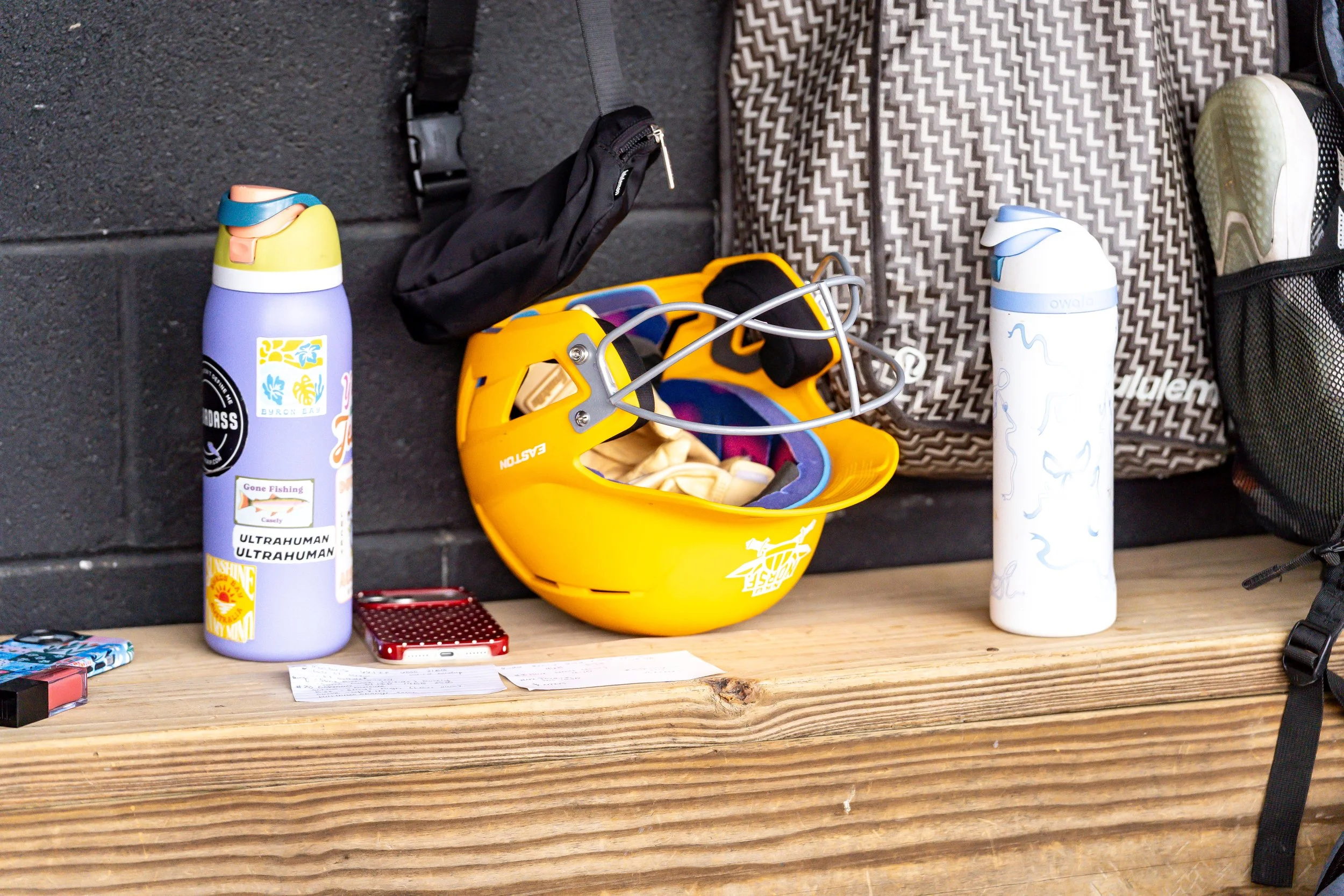 A wooden table with two water bottles, a small red box, a piece of paper, a yellow plastic sled, a black bag, and a patterned backpack against a brick wall.