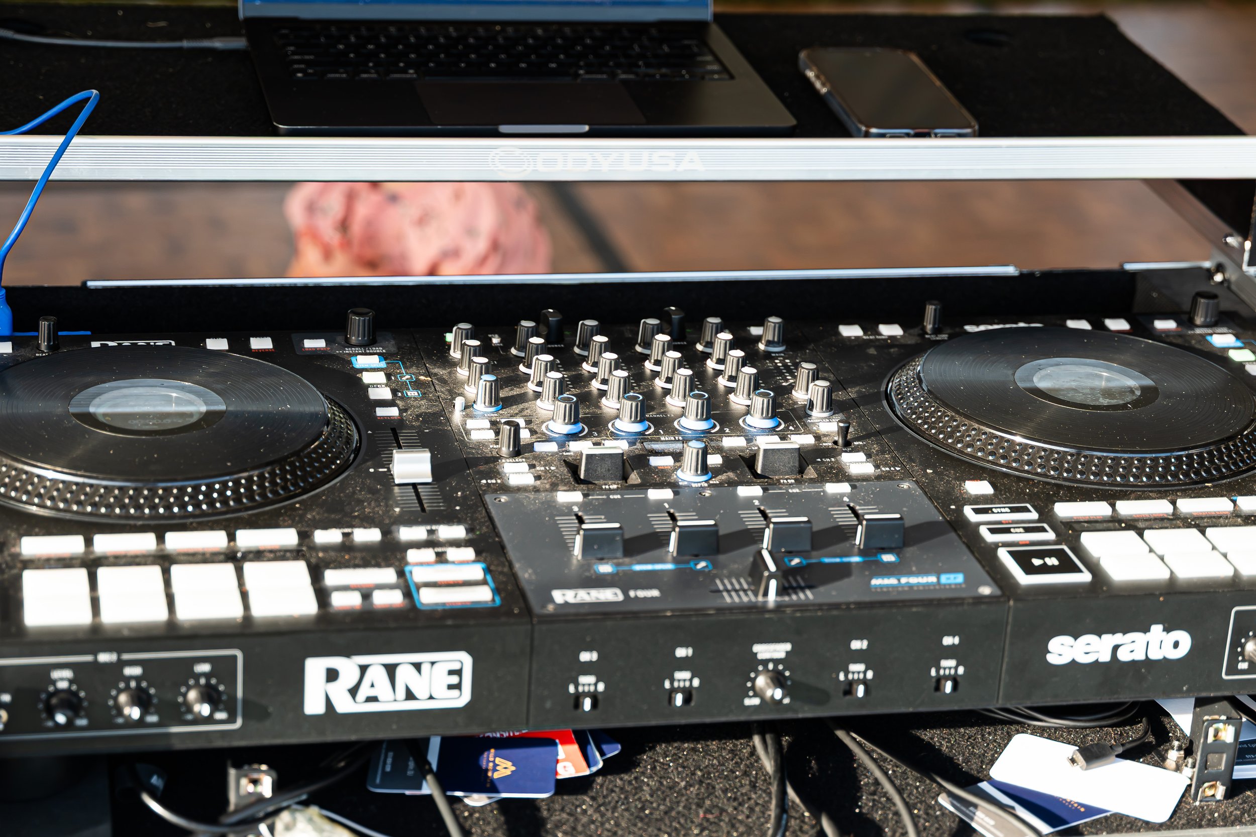 A DJ equipment setup on a black table, including a Rane mixer and a Serato DJ controller with numerous knobs and buttons, a laptop, and a smartphone. There are some items underneath the table, such as a credit card.