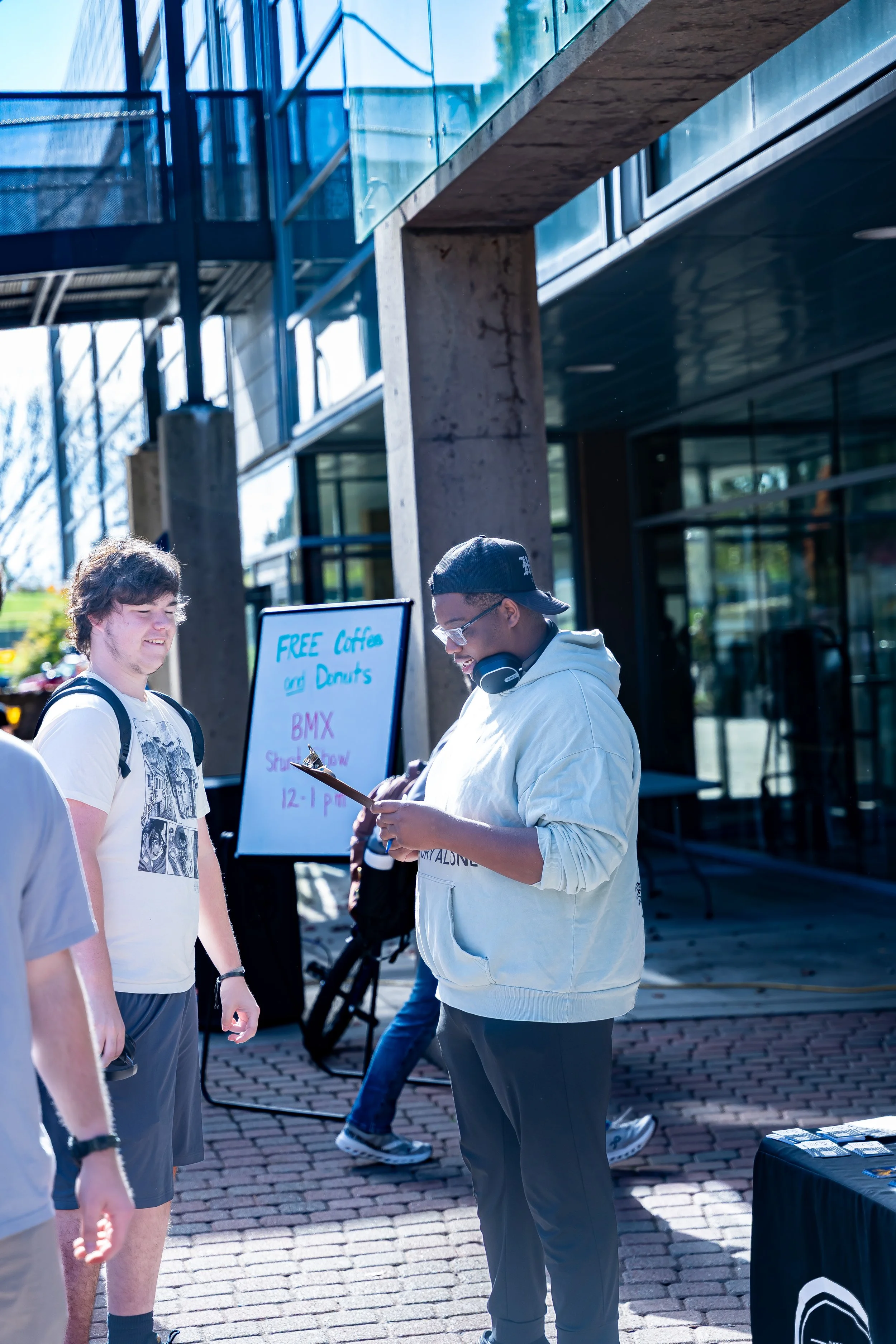 Two young men standing outdoors in front of a building, one with a backpack and the other wearing glasses and headphones around his neck, smiling at each other