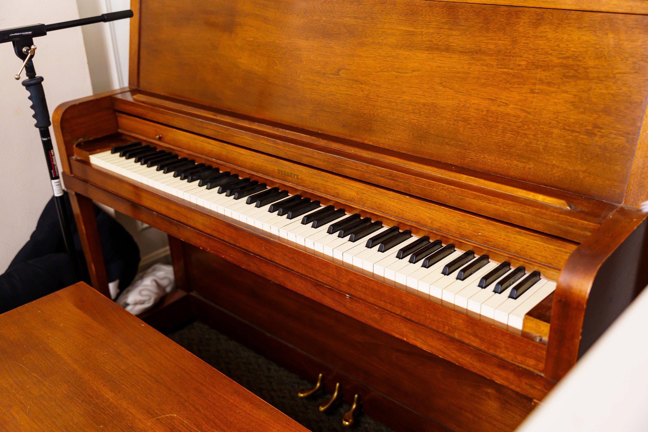 An upright wooden piano with black and white keys, positioned in a room with a white wall and a microphone stand nearby.