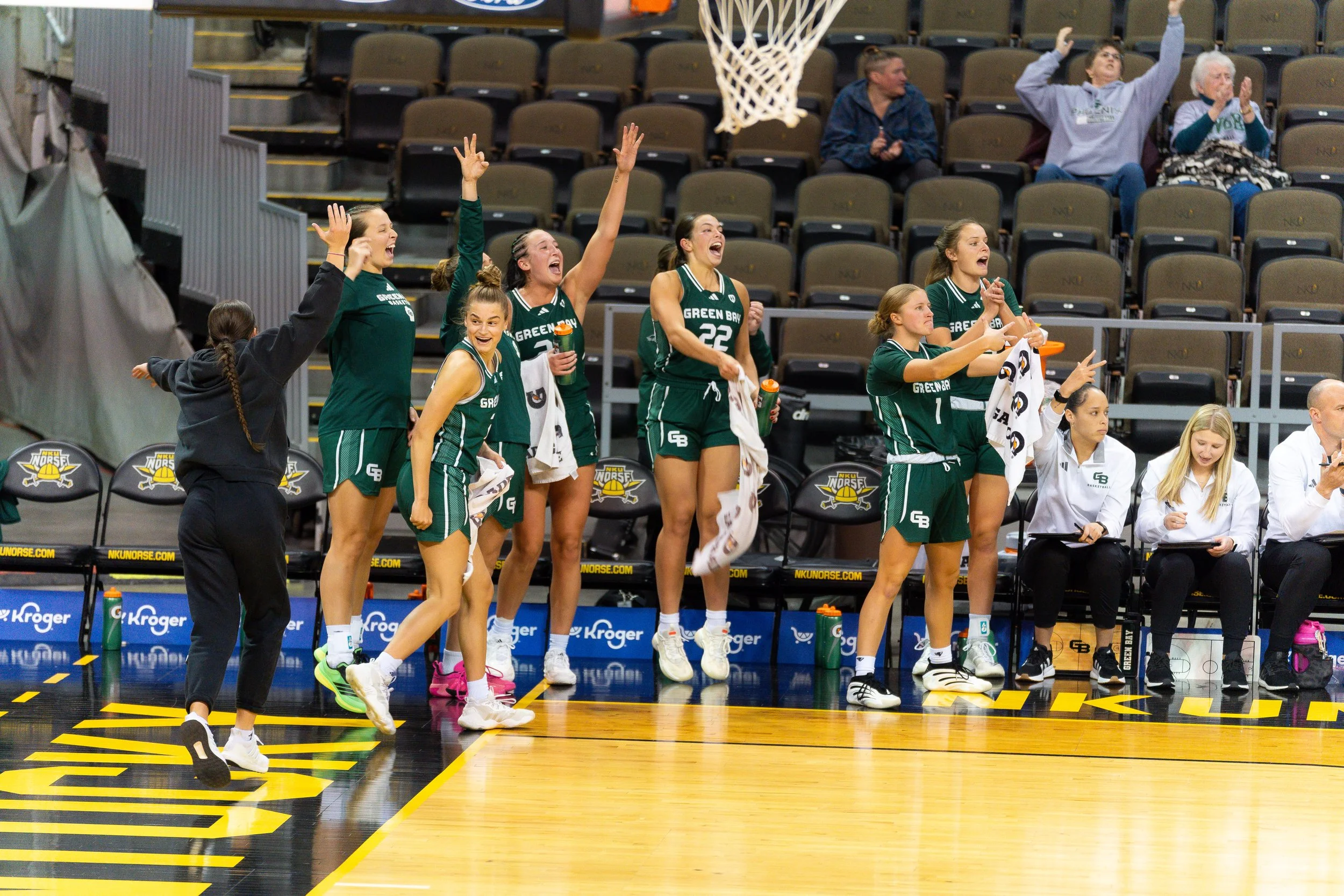 A women's basketball team in green uniforms on the bench, celebrating during a game. Some players are standing, cheering with arms raised, and holding towels. Others are sitting or standing near the bench, with a few clapping. The scene takes place i