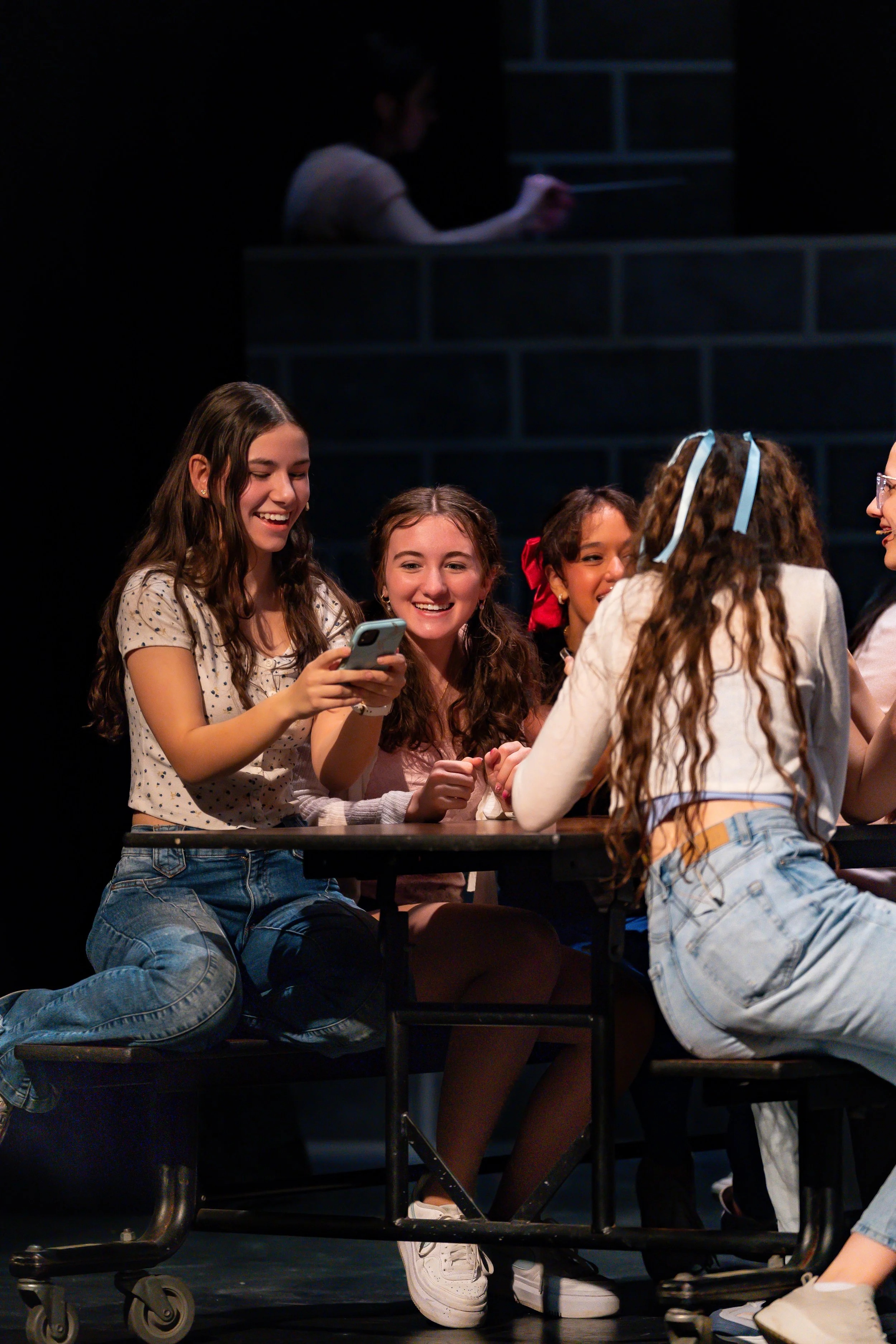 Group of young girls gathered around a table sharing a moment of laughter and conversation, with one girl looking at her phone.