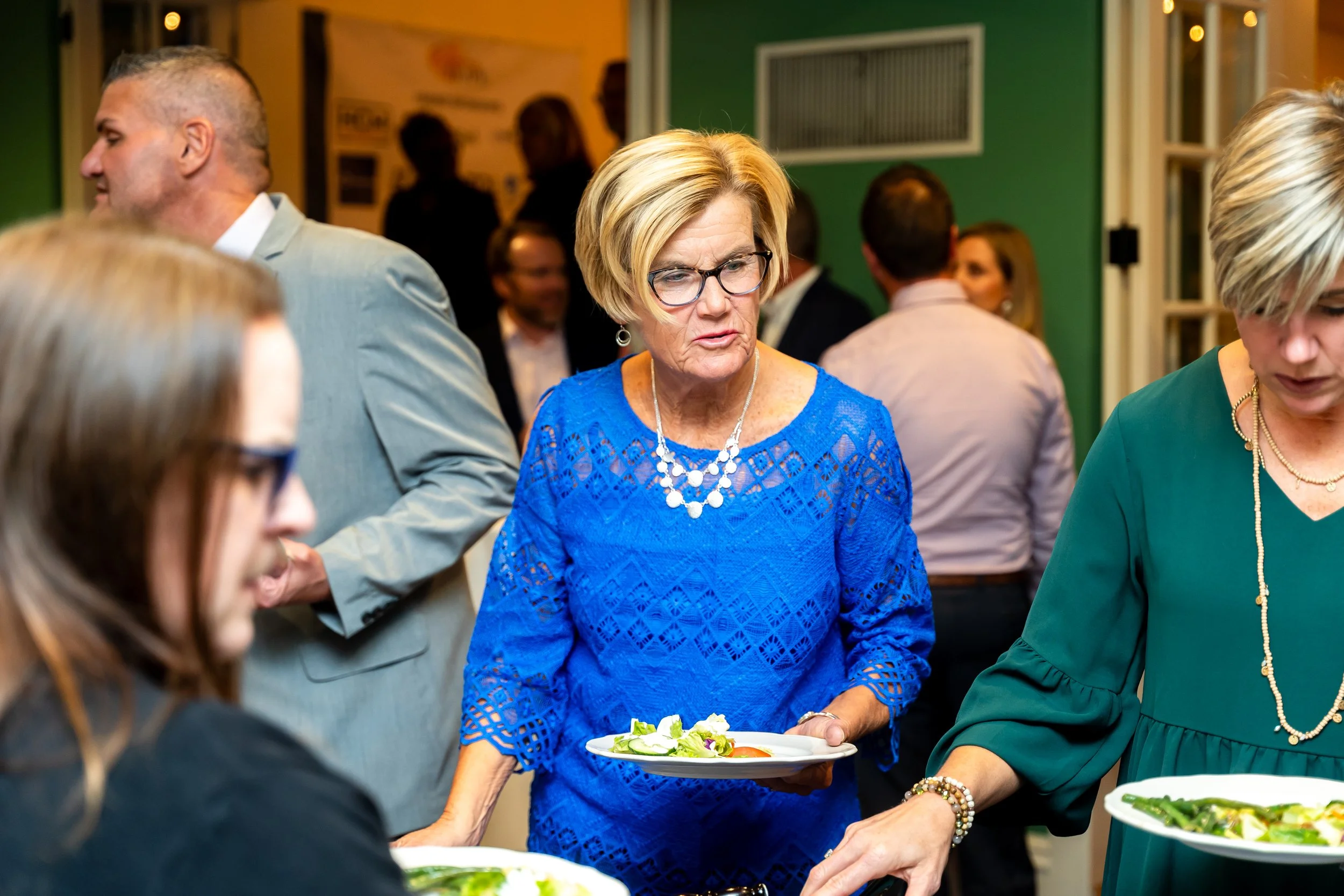 A woman with blonde hair, glasses, and a blue lace dress holding a plate of salad in a social gathering.