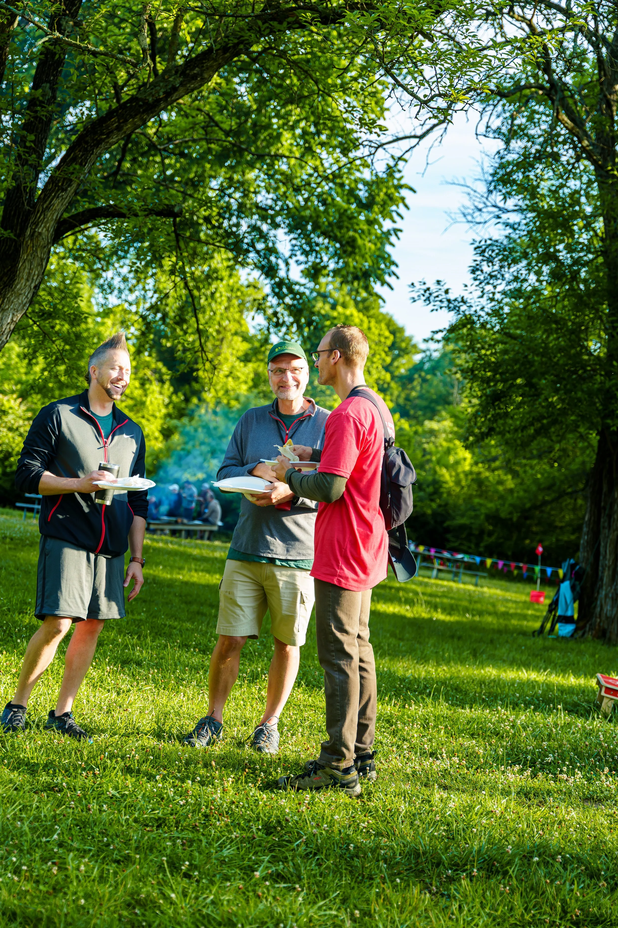 Three men standing and talking outdoors in a park with green grass and tall trees, smiling and holding plates of food, with a picnic area and colorful flags in the background.