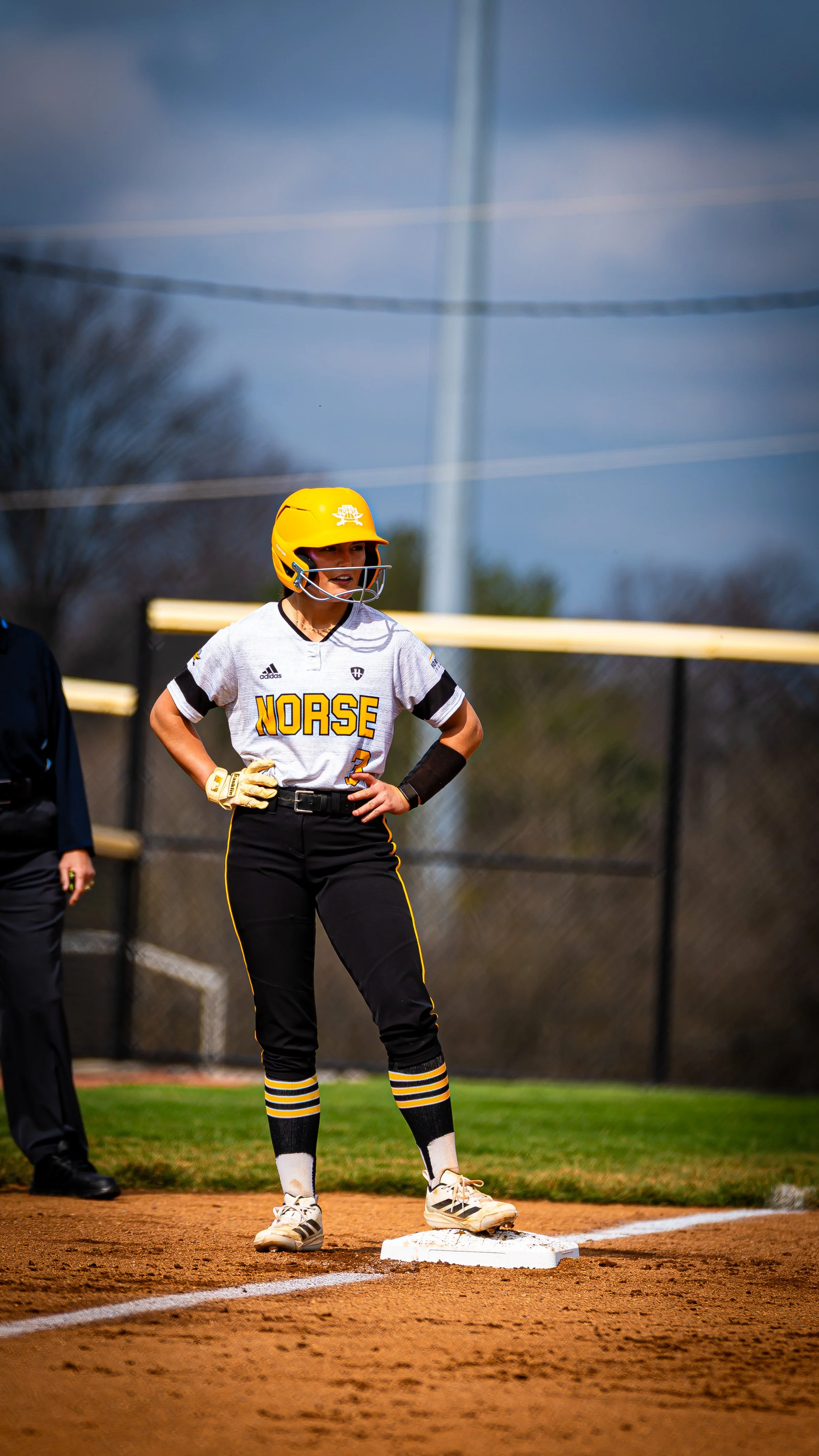 A female softball player wearing a yellow helmet and uniform with 'Norse' printed on her shirt, standing on a base during a game.