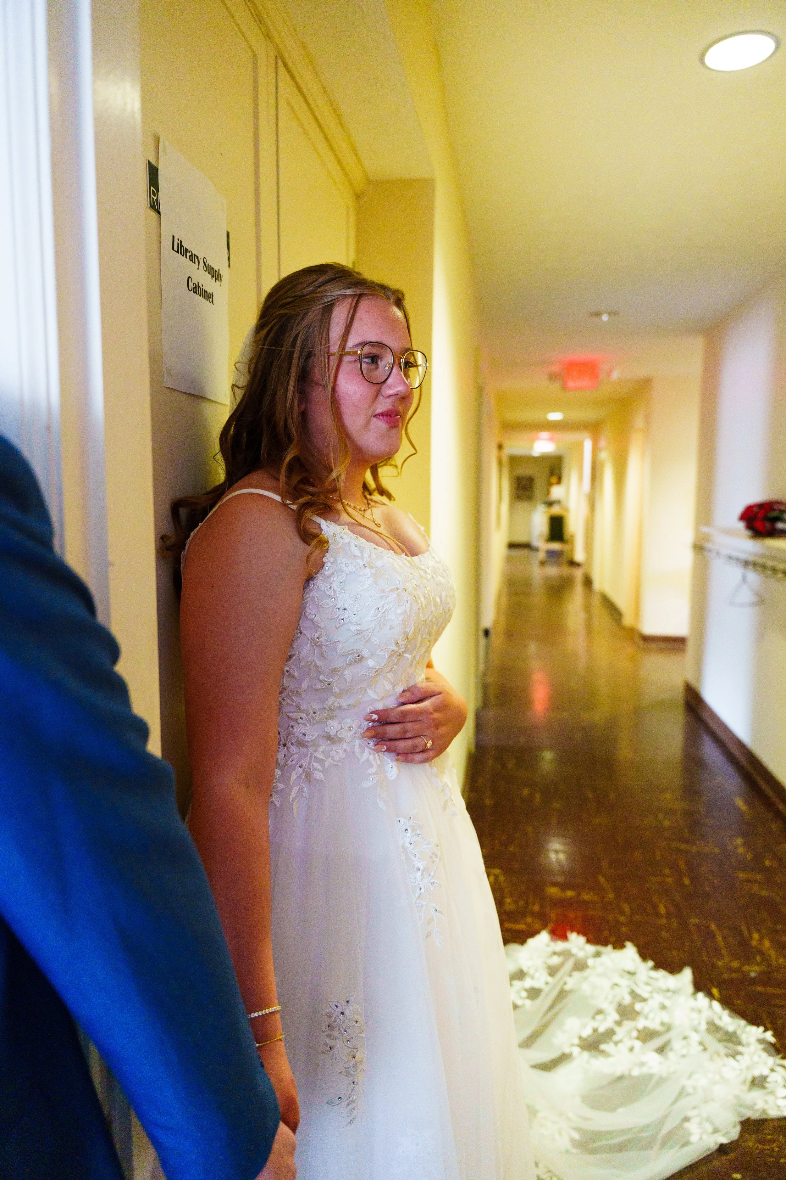 A woman in a wedding dress standing in a hallway next to a person in a blue suit, with a sign on the wall reading 'Library Supply Cabinet'.