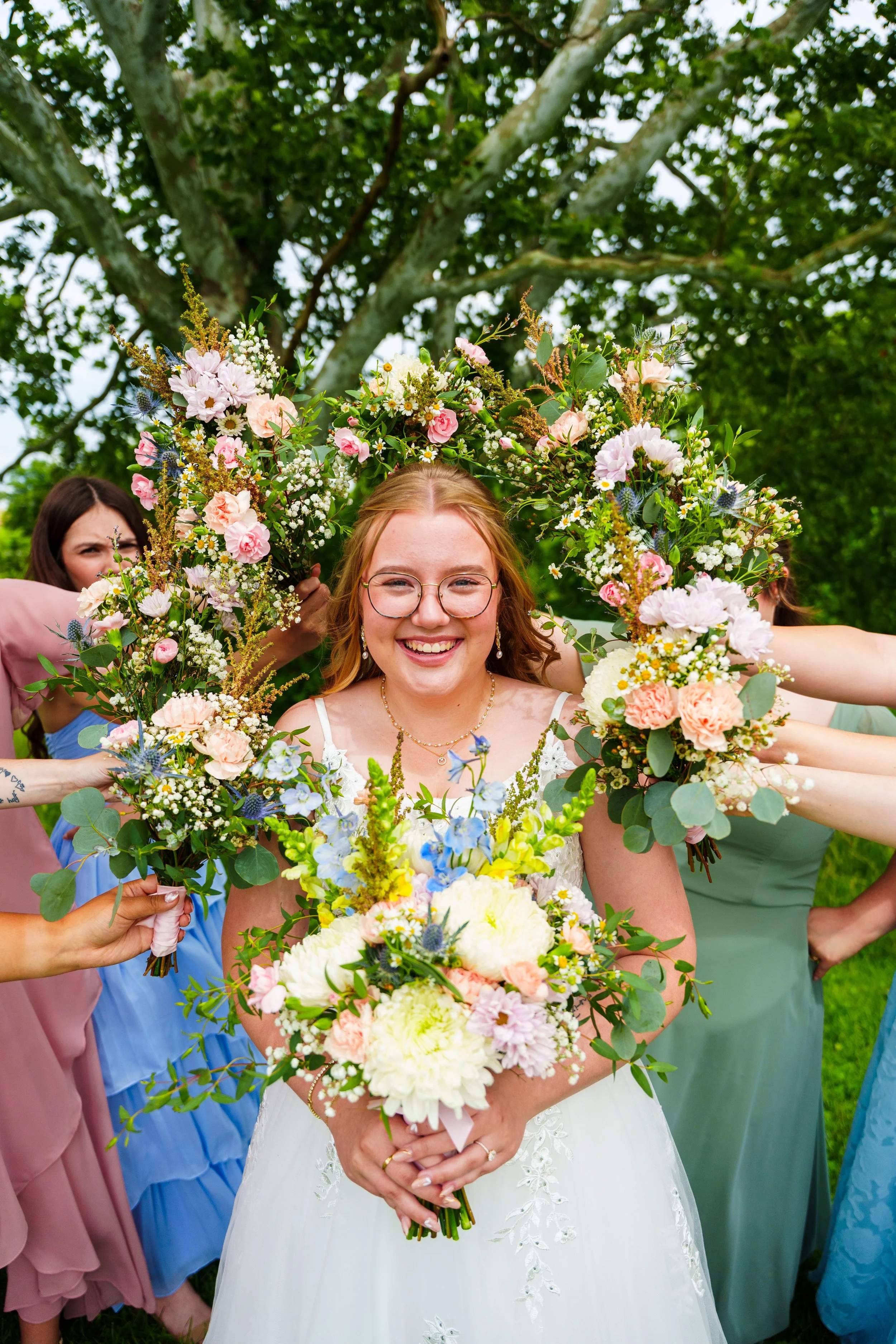 A smiling bride with glasses holding a large bouquet of white and pastel pink flowers, surrounded by friends in colorful dresses during an outdoor wedding.