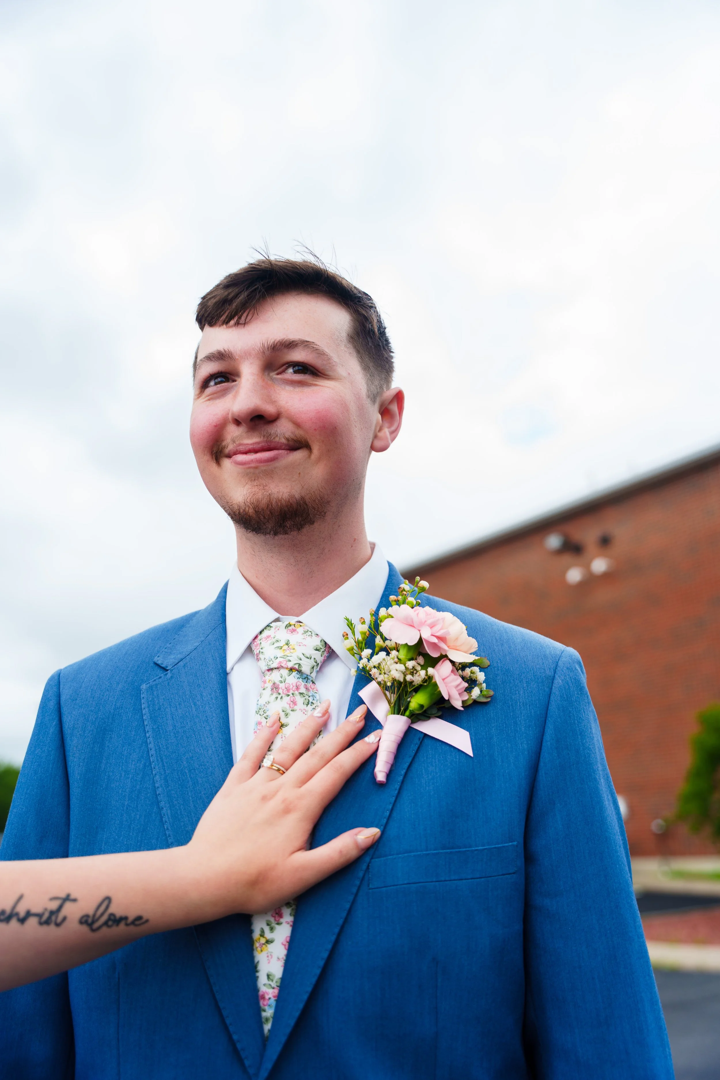 A young man in a blue suit holding a pink and white boutonniere, with a hand resting on his chest, standing outdoors against a cloudy sky and brick building background.