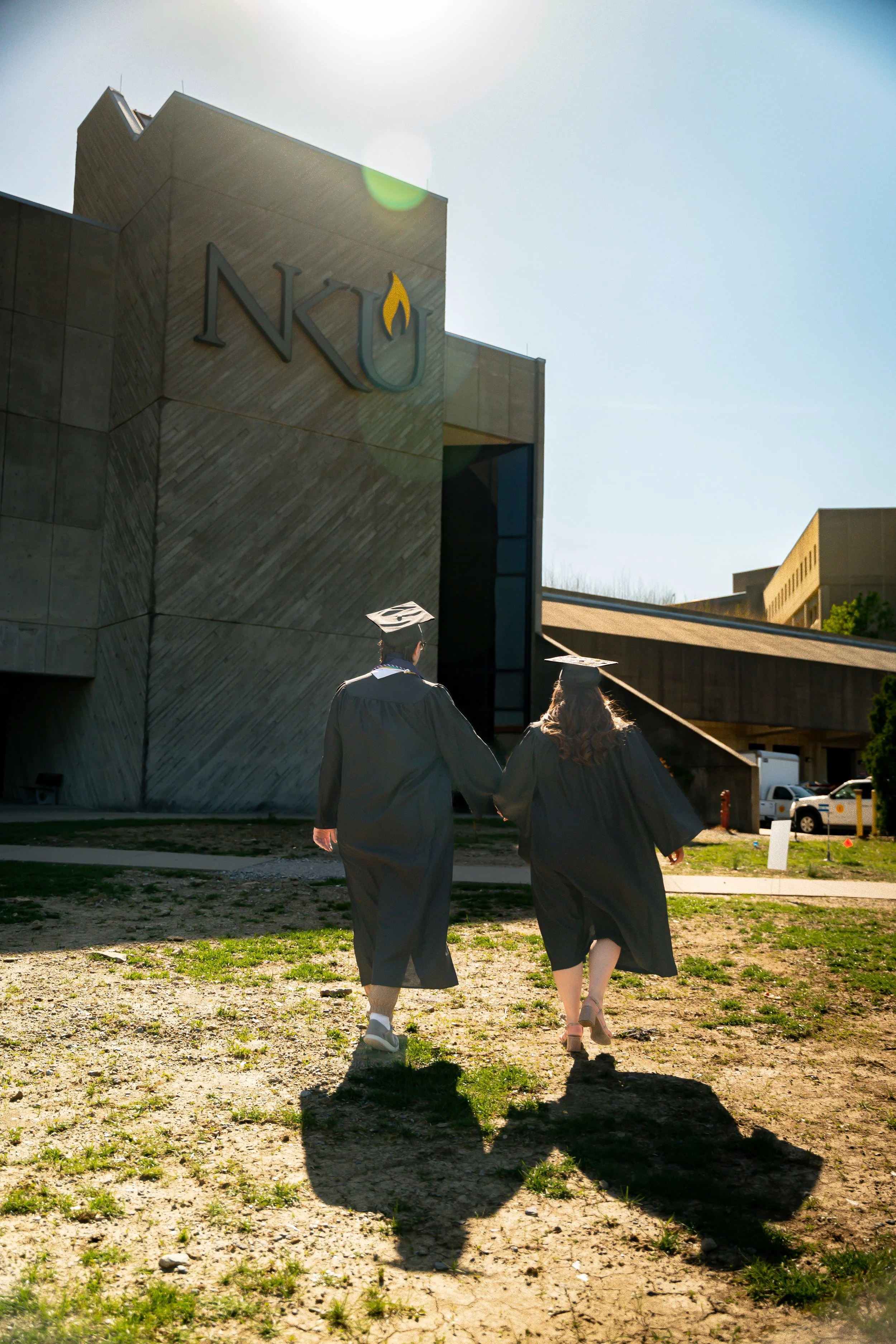 Two graduates wearing black caps and gowns walking away from the camera towards a modern university building with the Nku logo on it, casting long shadows on the ground.