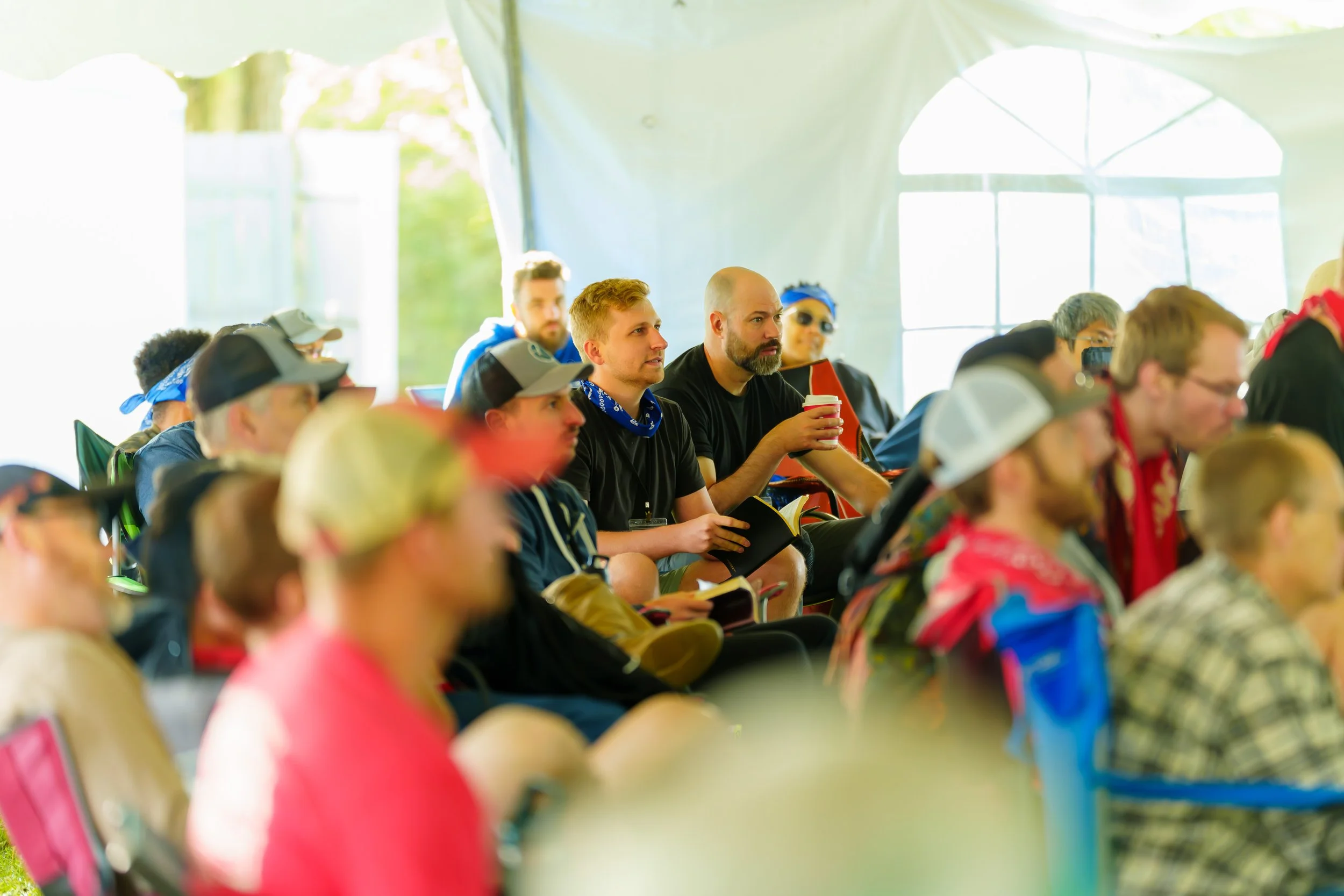 A group of people sitting inside a large tent, attentively listening, with some holding drinks and notebooks, during an outdoor event or conference.
