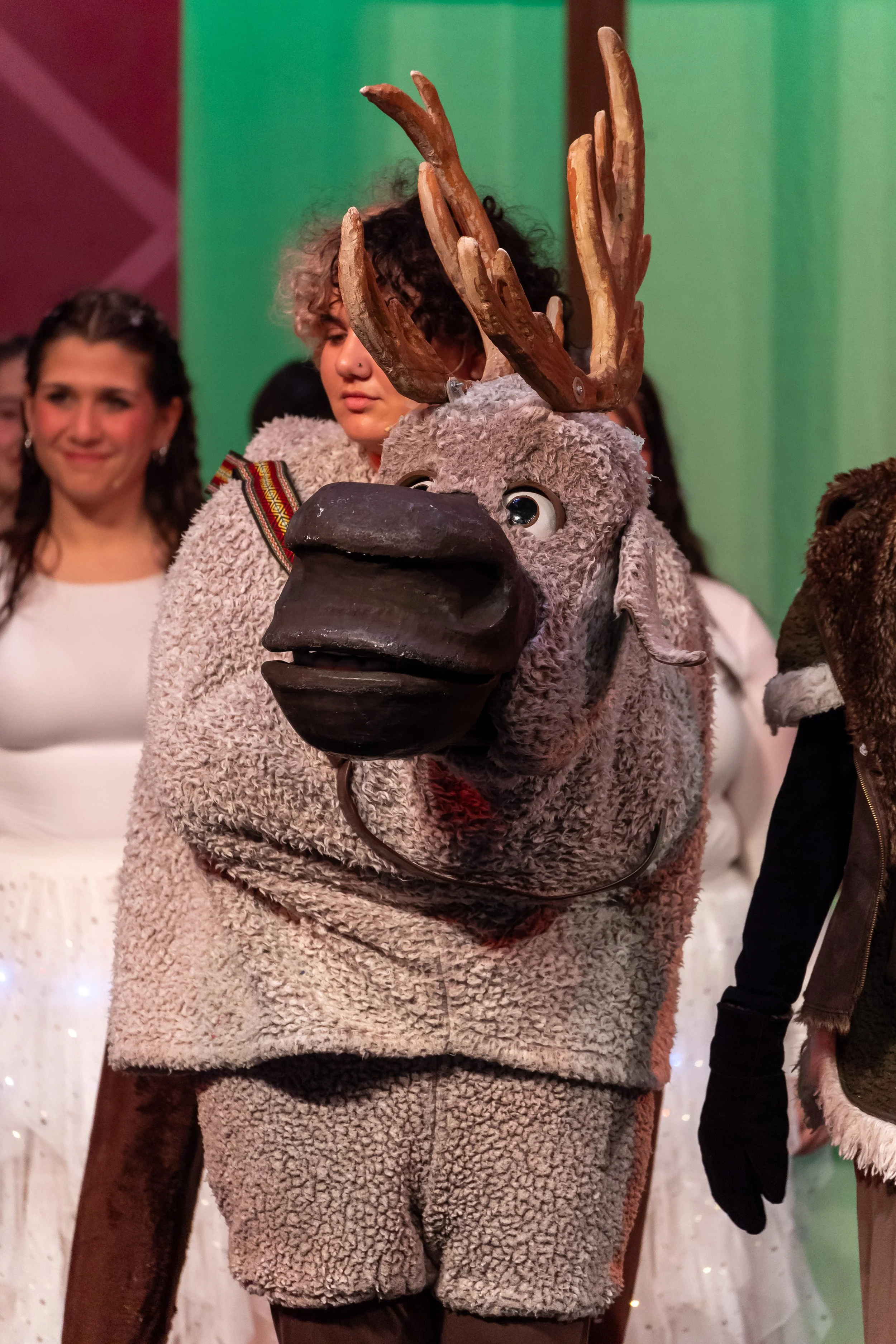 Person in a plush, teddy bear costume with large antlers on its head, standing indoors at a festive event with people in background.