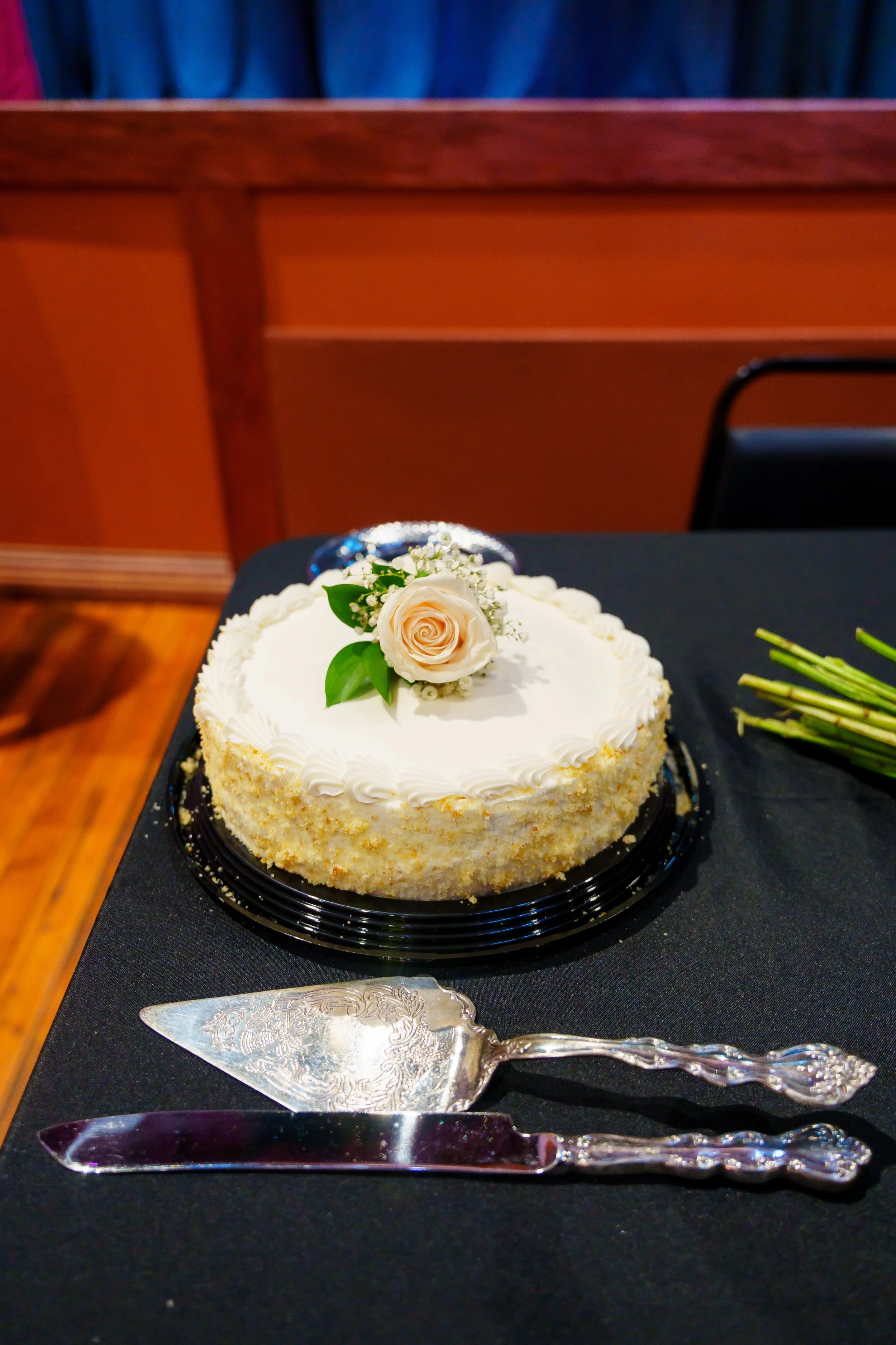 A round white cake decorated with a cream rose and small white flowers, placed on a black table with silver cake utensils nearby. In the background, an orange wall and blue curtains are visible.