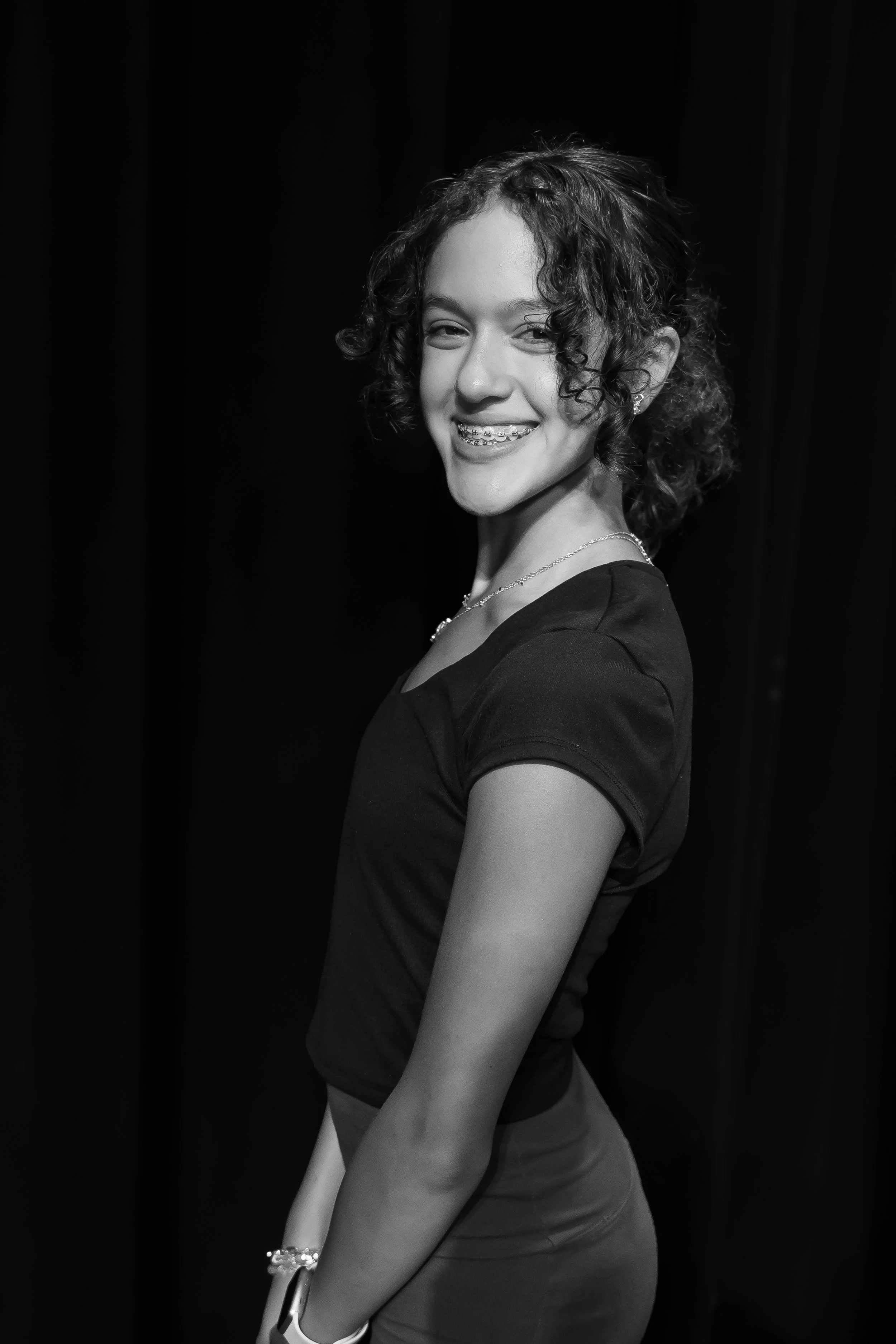 A young woman with curly hair smiling, wearing a black top, earrings, and a necklace, standing against a dark background.