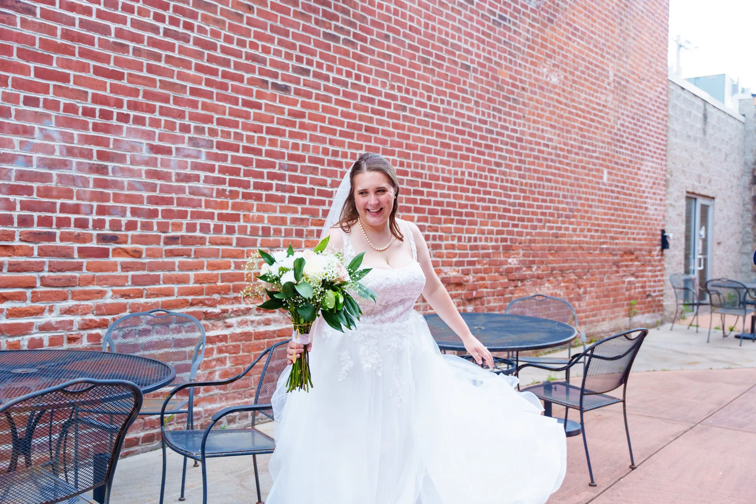 A smiling bride in a white wedding gown holding a bouquet of white flowers and greenery, standing in front of a red brick wall on an outdoor patio with black metal chairs and tables.