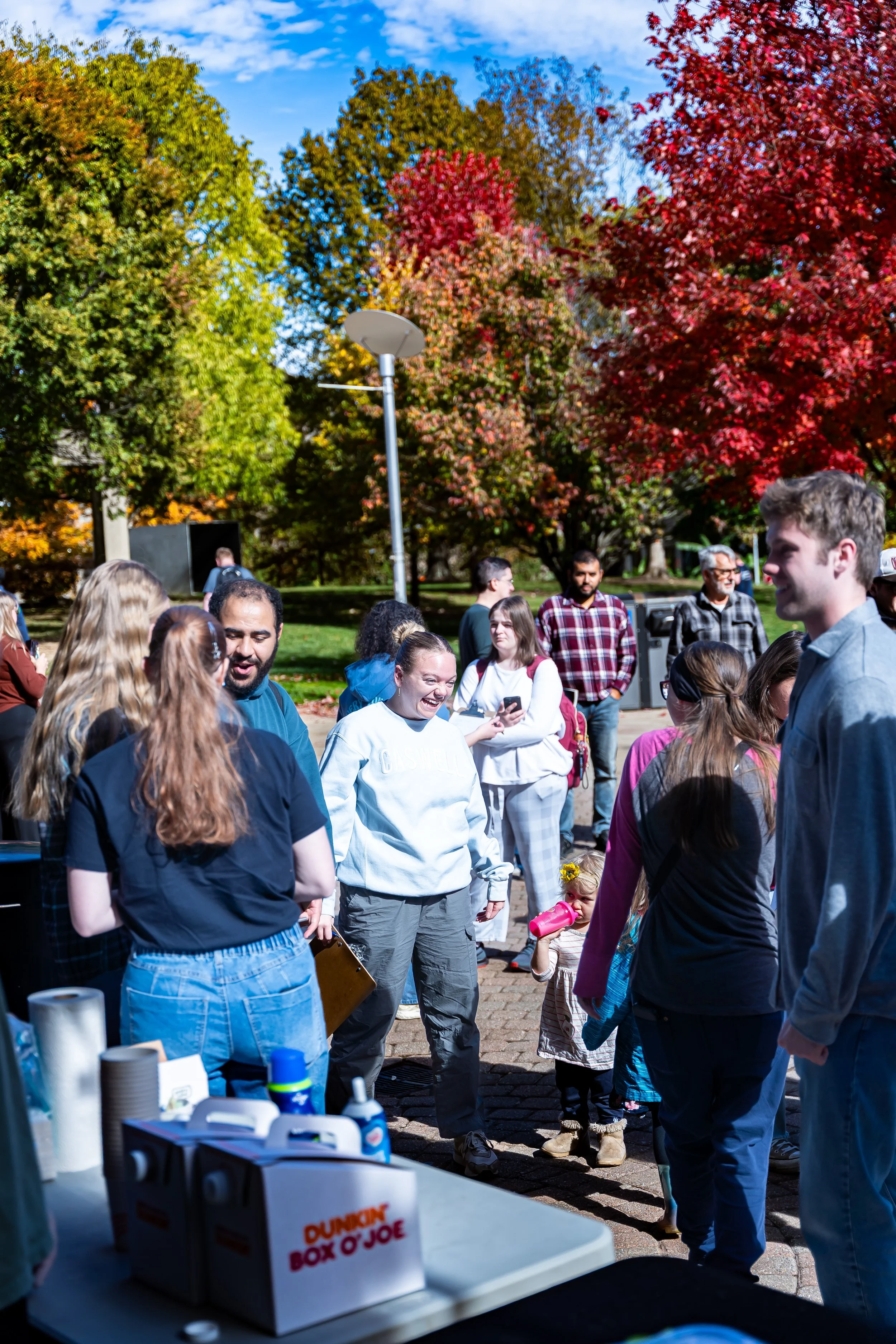 A group of people gathered outdoors on a sunny day, with autumn-colored trees in the background. Some individuals are smiling and talking, while a little girl with a pink water bottle is standing among adults. A table in the foreground holds various 