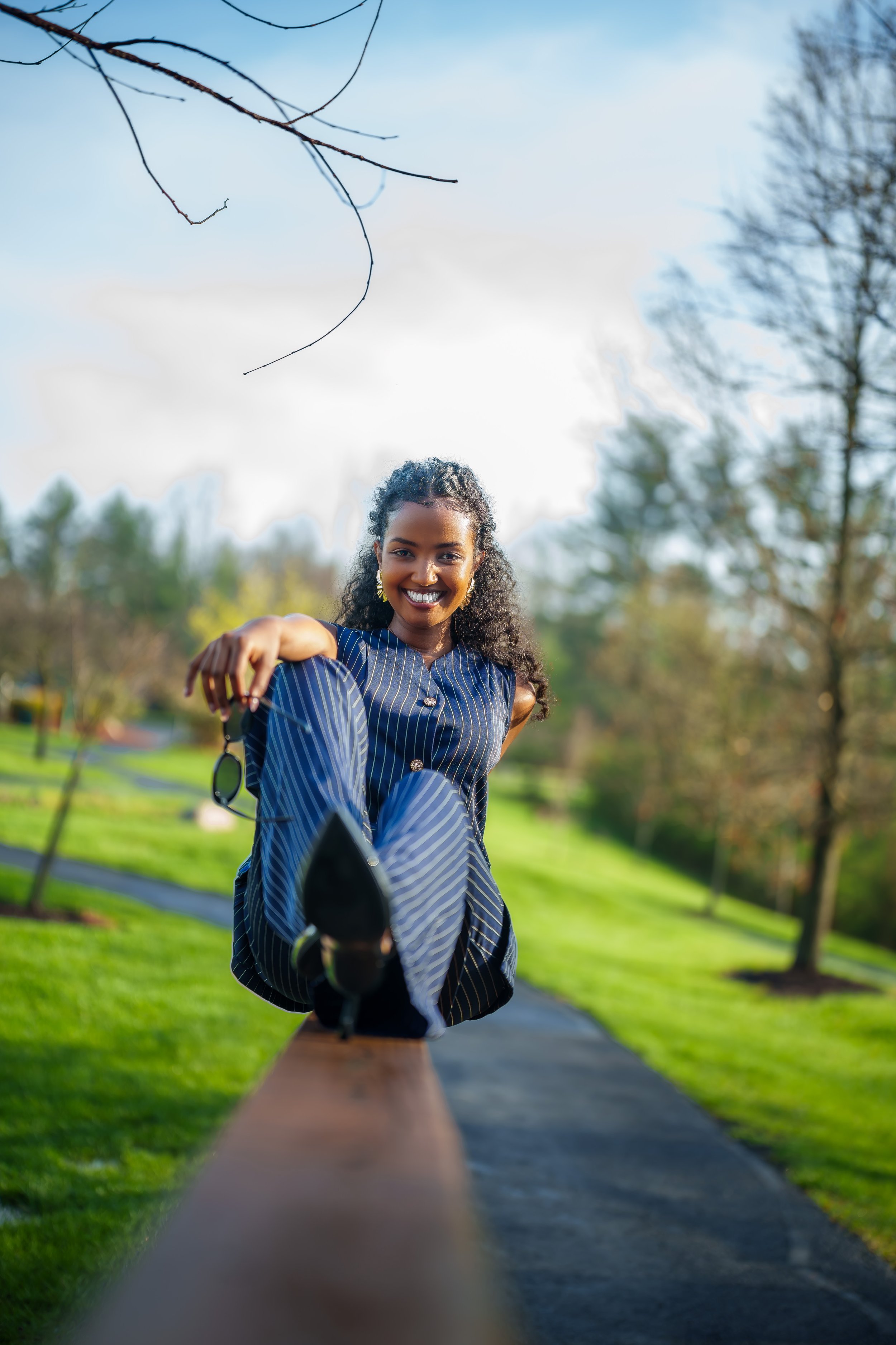 Young woman smiling and stretching her leg on a park bench outdoors on a sunny day with trees and a walking path in the background.