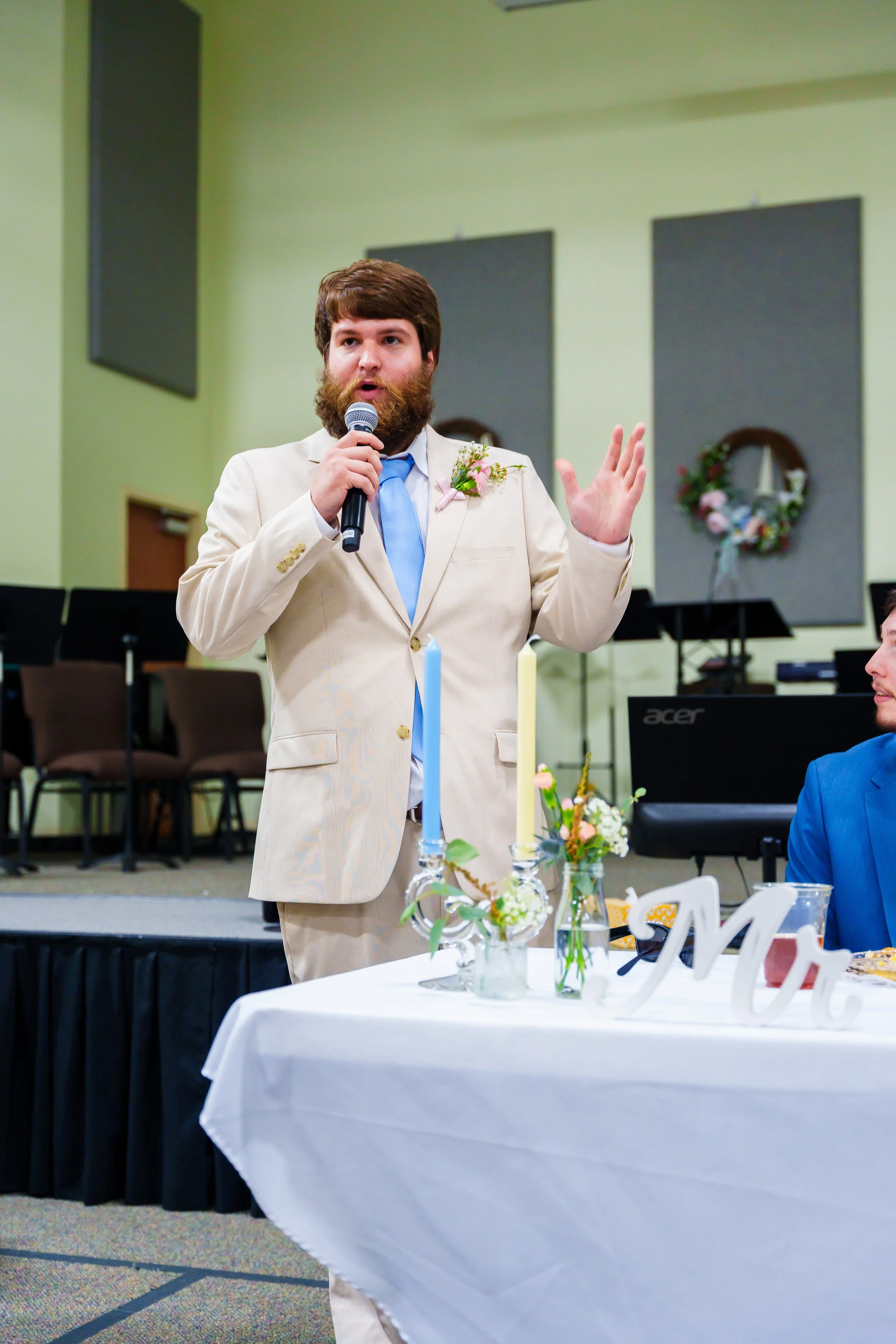 A man with a beard and brown hair speaking into a microphone at a wedding reception, wearing a beige suit with a light blue tie and a pink flower boutonniere. There are candles, flowers, and wedding decorations on the table in front of him.