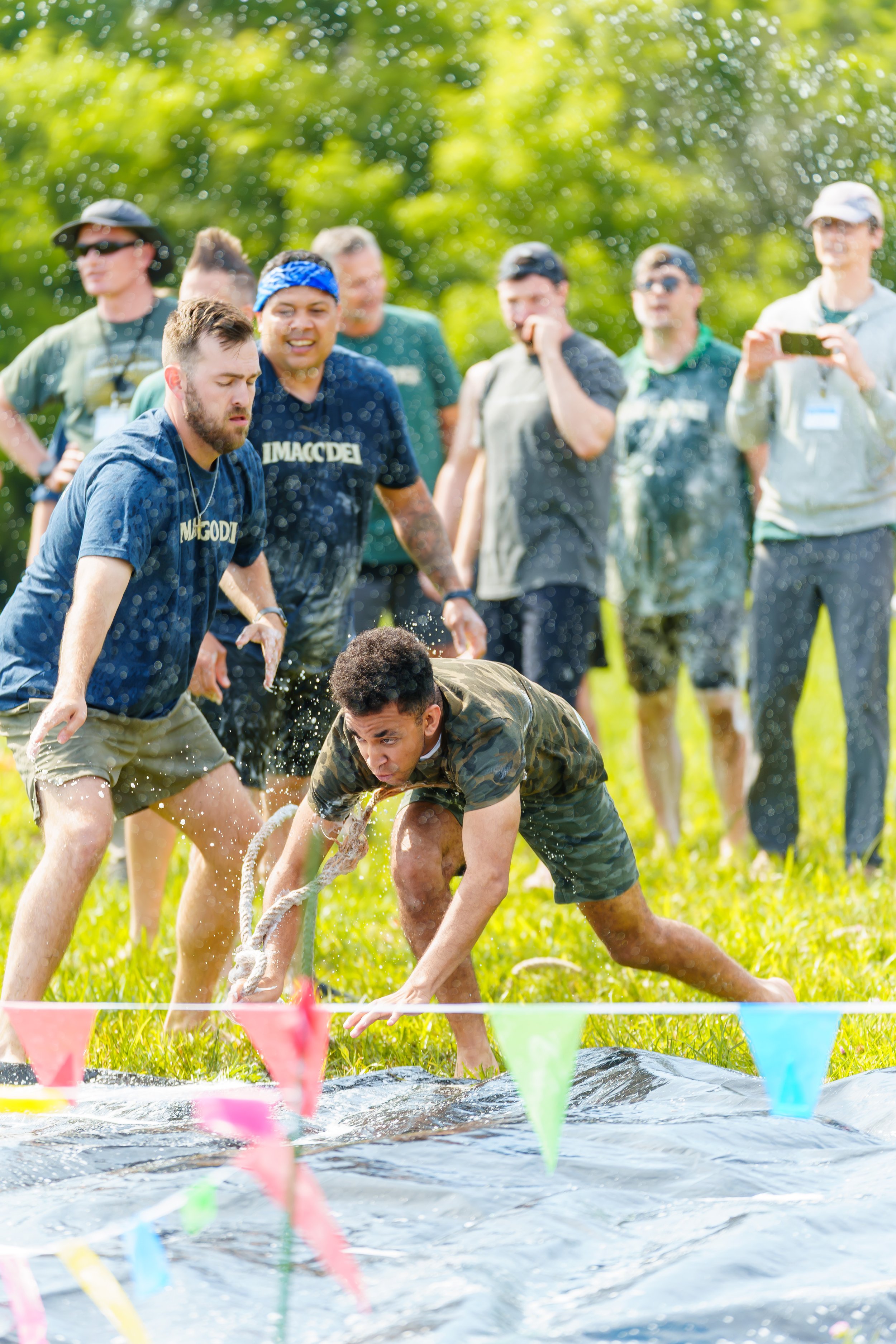 Group of people participating in an outdoor water activity, some are getting wet while others are watching, with a grassy field and trees in the background.