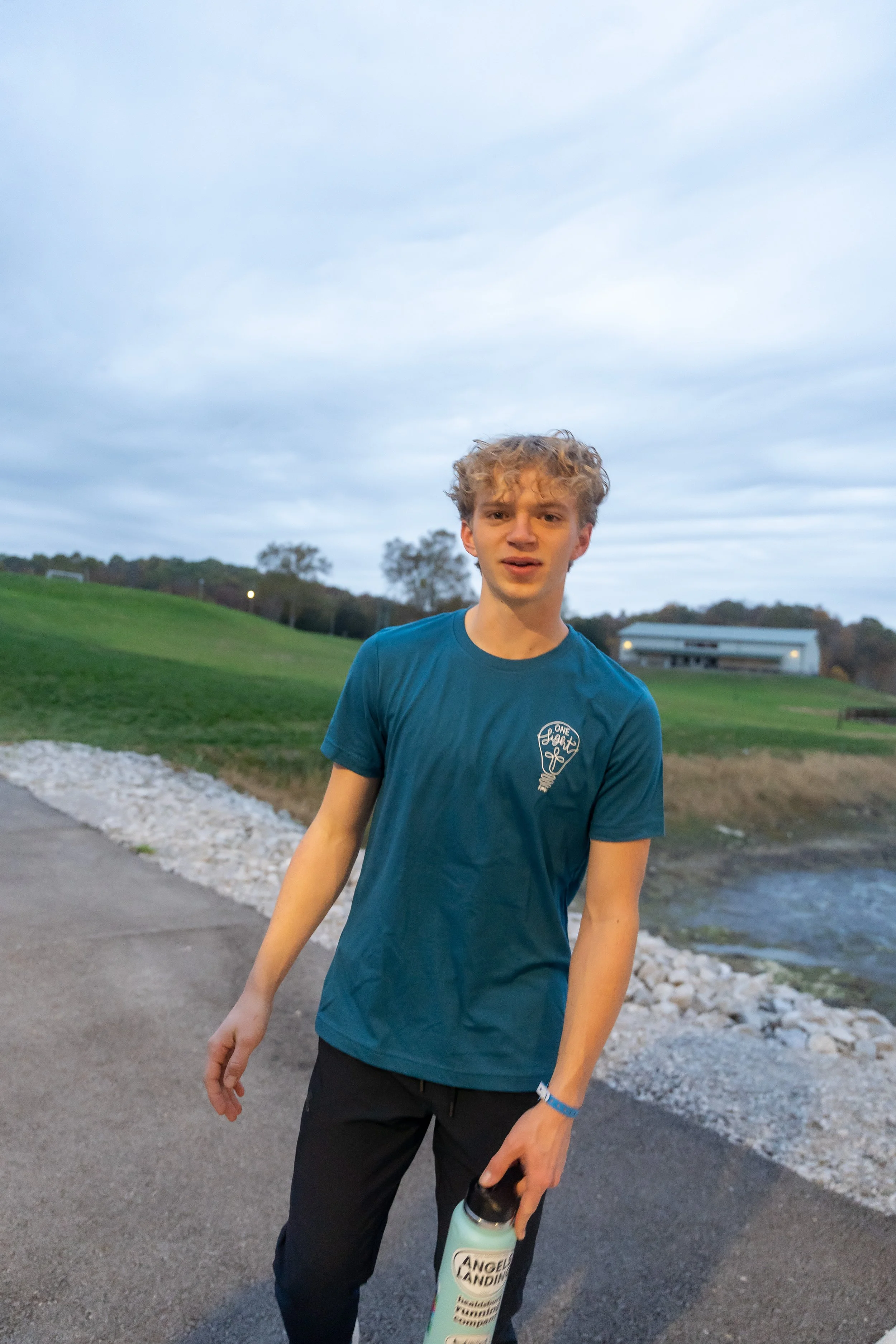 A young man standing outdoors near a water body, holding a water bottle, wearing a blue t-shirt with a light bulb logo, black pants, and a blue wristband. The background features a grassy hill, trees, a building, and an overcast sky.
