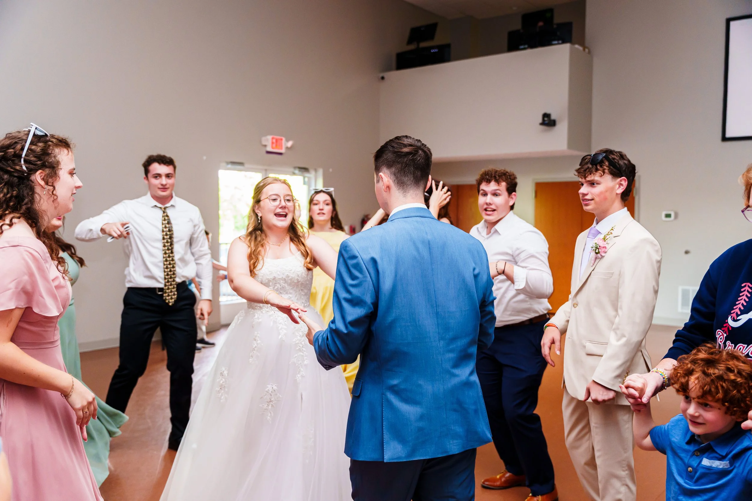 Group of young people dancing and celebrating at a wedding reception indoors, with a bride and groom in the center.