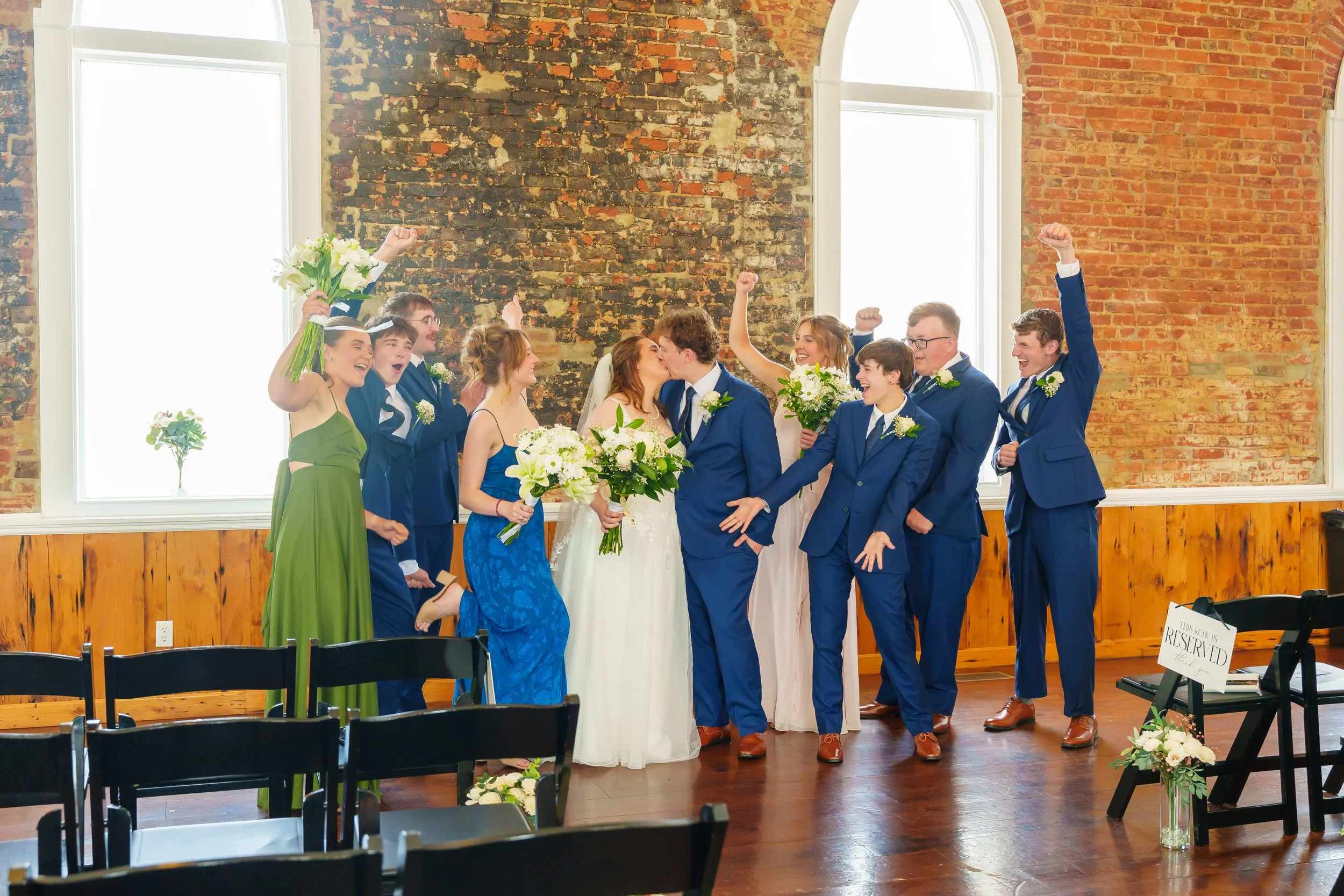 A group of wedding guests celebrating, with some cheering and raising their fists, while a bride and groom kiss in the center, in a room with brick walls and large windows.
