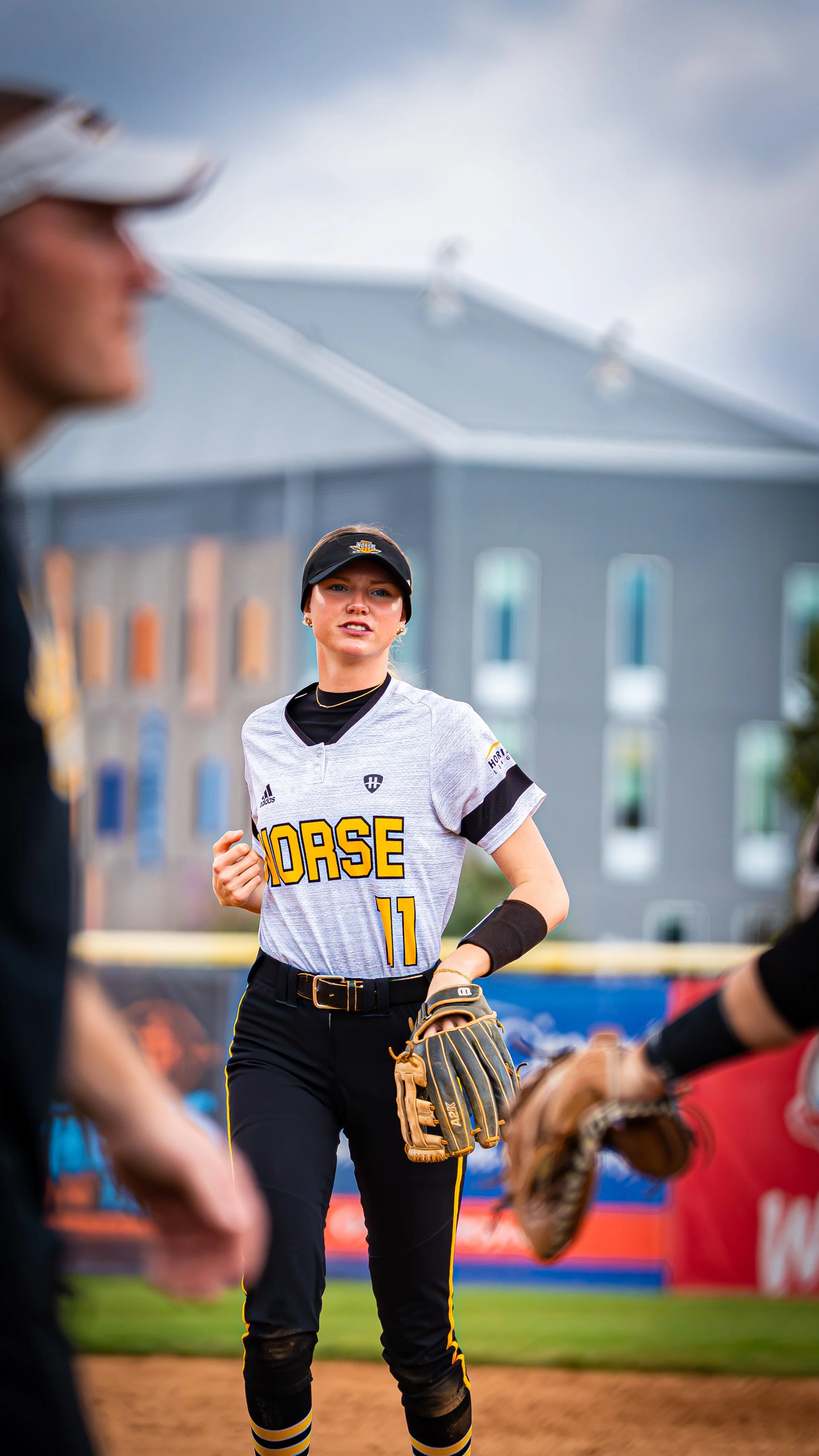 A female baseball player in a white uniform with black and yellow accents, wearing a cap and glove, standing on a baseball field during a game. She appears to be engaged in a conversation with players around her.