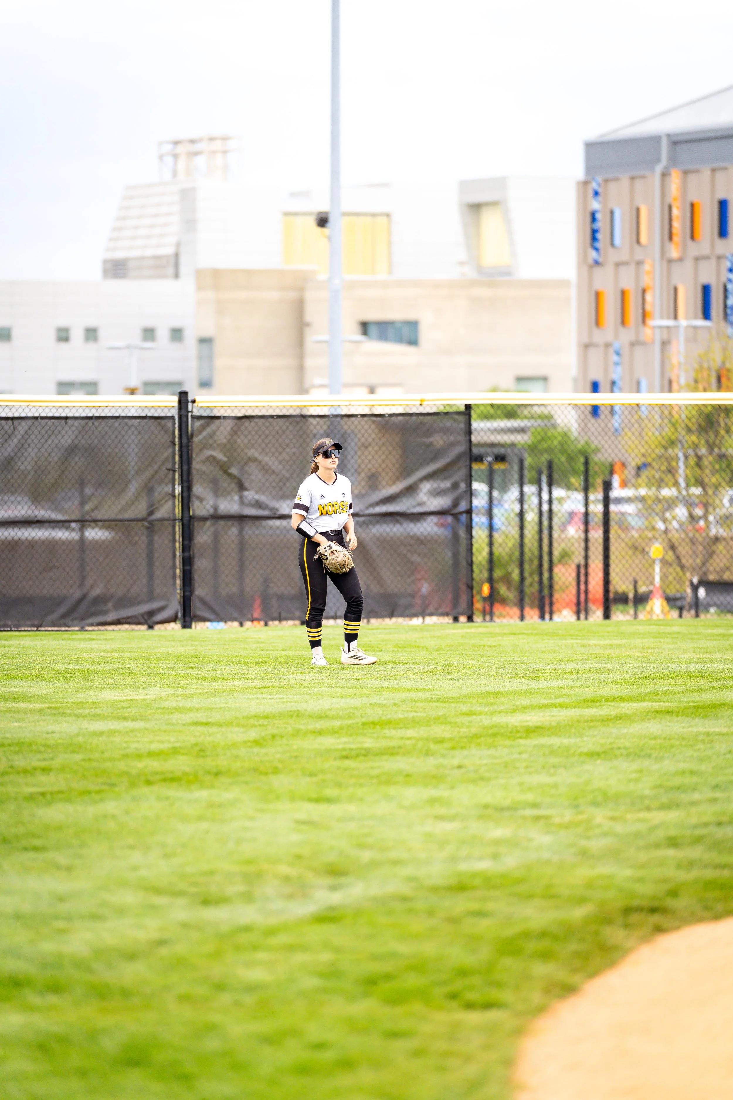 A female softball player dressed in a black and yellow uniform, standing on the field with a glove, behind a black fence with a yellow top, in an urban park or sports complex with buildings in the background.