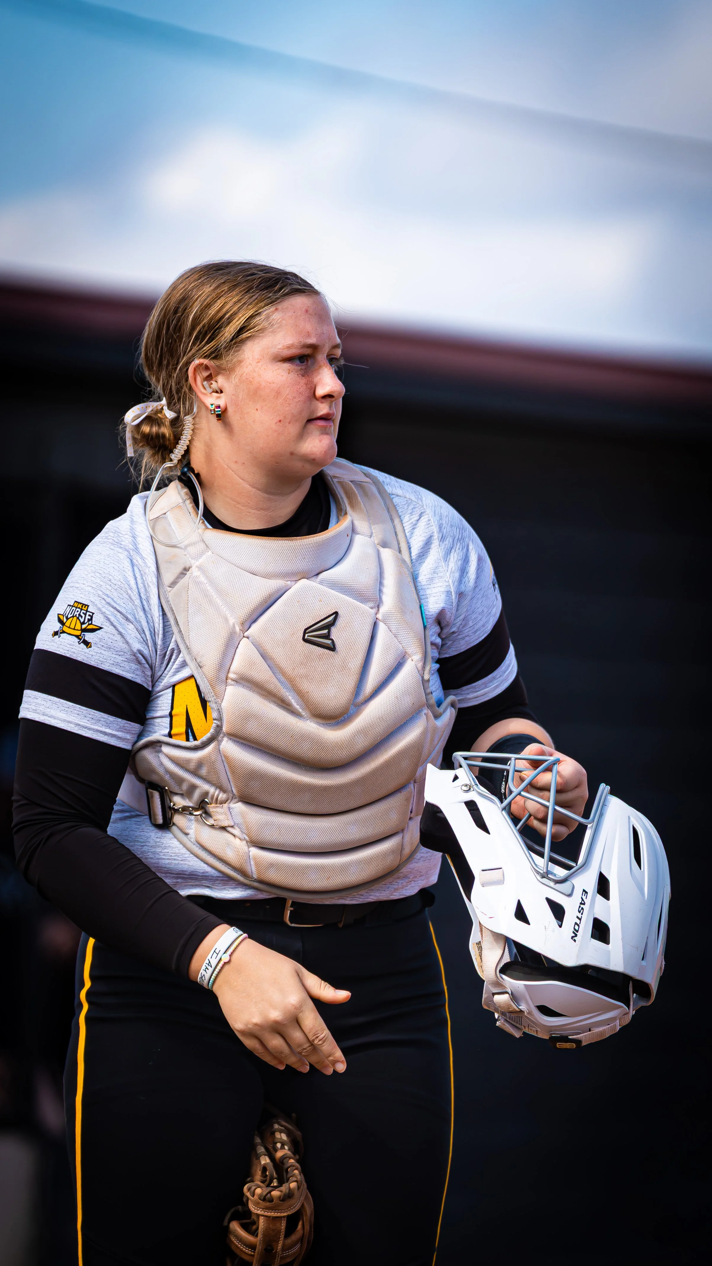 A young female softball catcher wearing a beige chest protector, holding a white Easton helmet with a face guard, and dressed in black athletic clothing with yellow piping.
