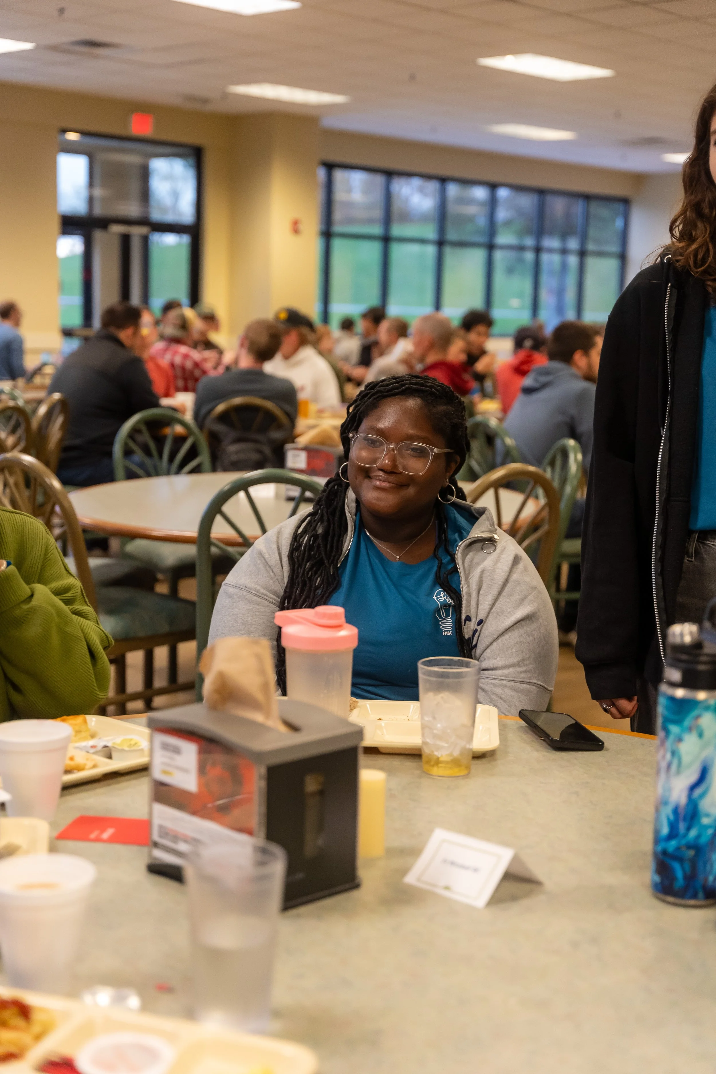 Young woman with glasses and long hair seated at a cafeteria table, surrounded by plates, cups, and food. People are in the background, sitting and socializing in a well-lit room with large windows.