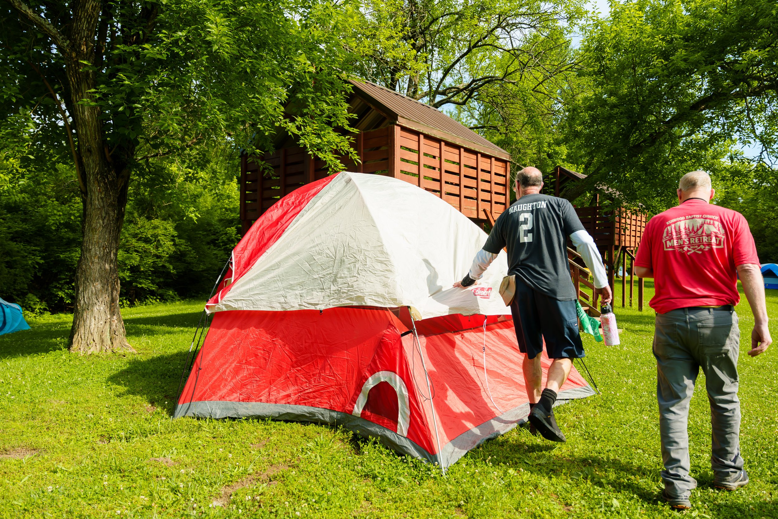 Two men setting up a red and white tent outdoors in a grassy area surrounded by trees, with wooden structures in the background.
