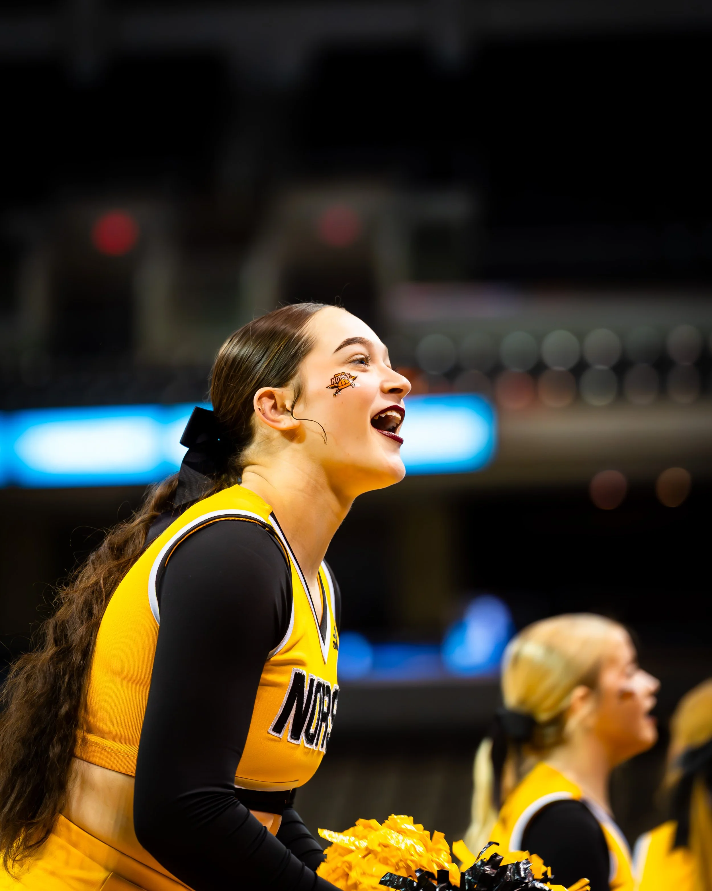Cheerleader with long curly hair, yellow and black uniform, smiling with mouth open, holding yellow pom-poms, at a sports event with a blurred audience and scoreboard in the background.