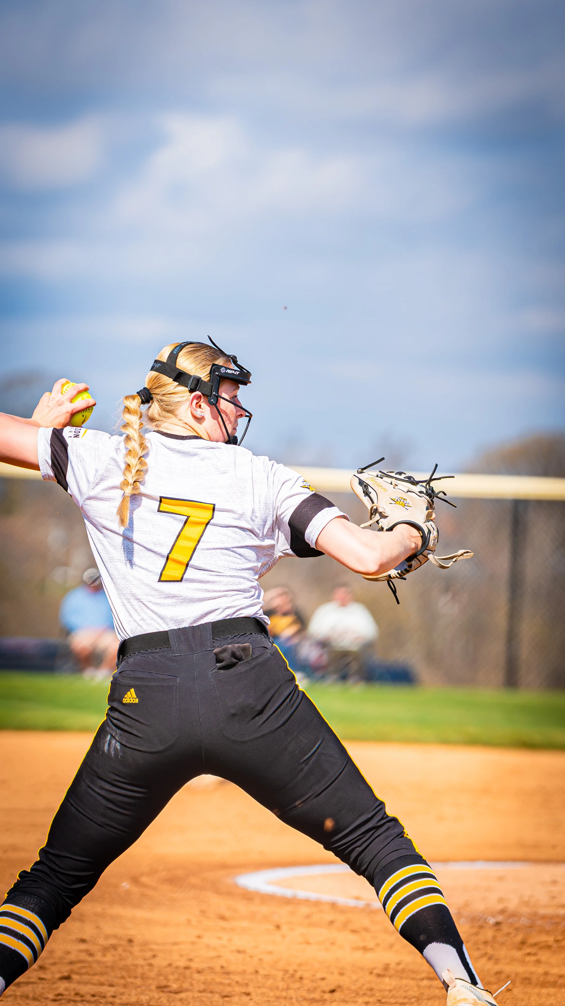 A female softball player in uniform with the number 7 on her back, in a pitching stance on the field, preparing to throw a pitch.