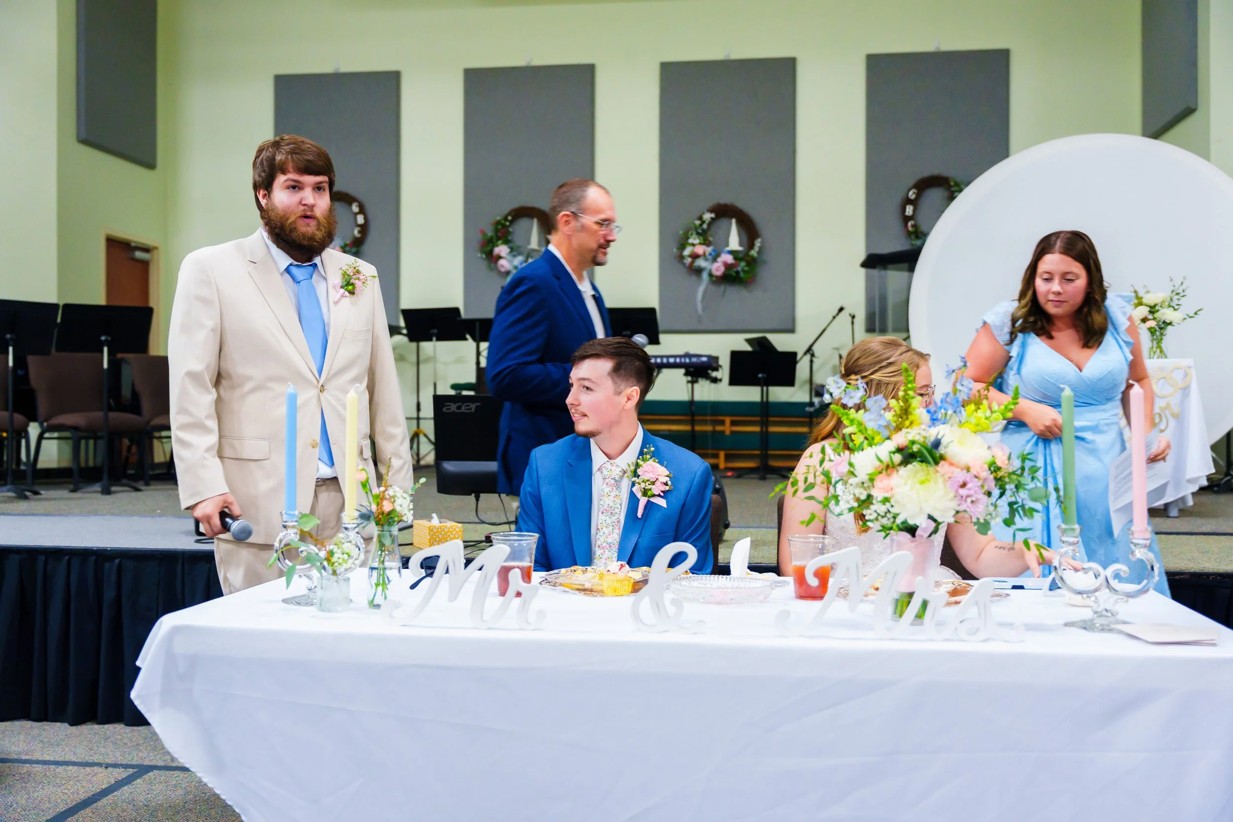 A wedding celebration in a banquet hall with floral decorations and a cake. The groom is seated wearing a blue suit, while the bride with flowers in her hair is reaching over the table. Two men are standing nearby, one in a beige suit and another in 