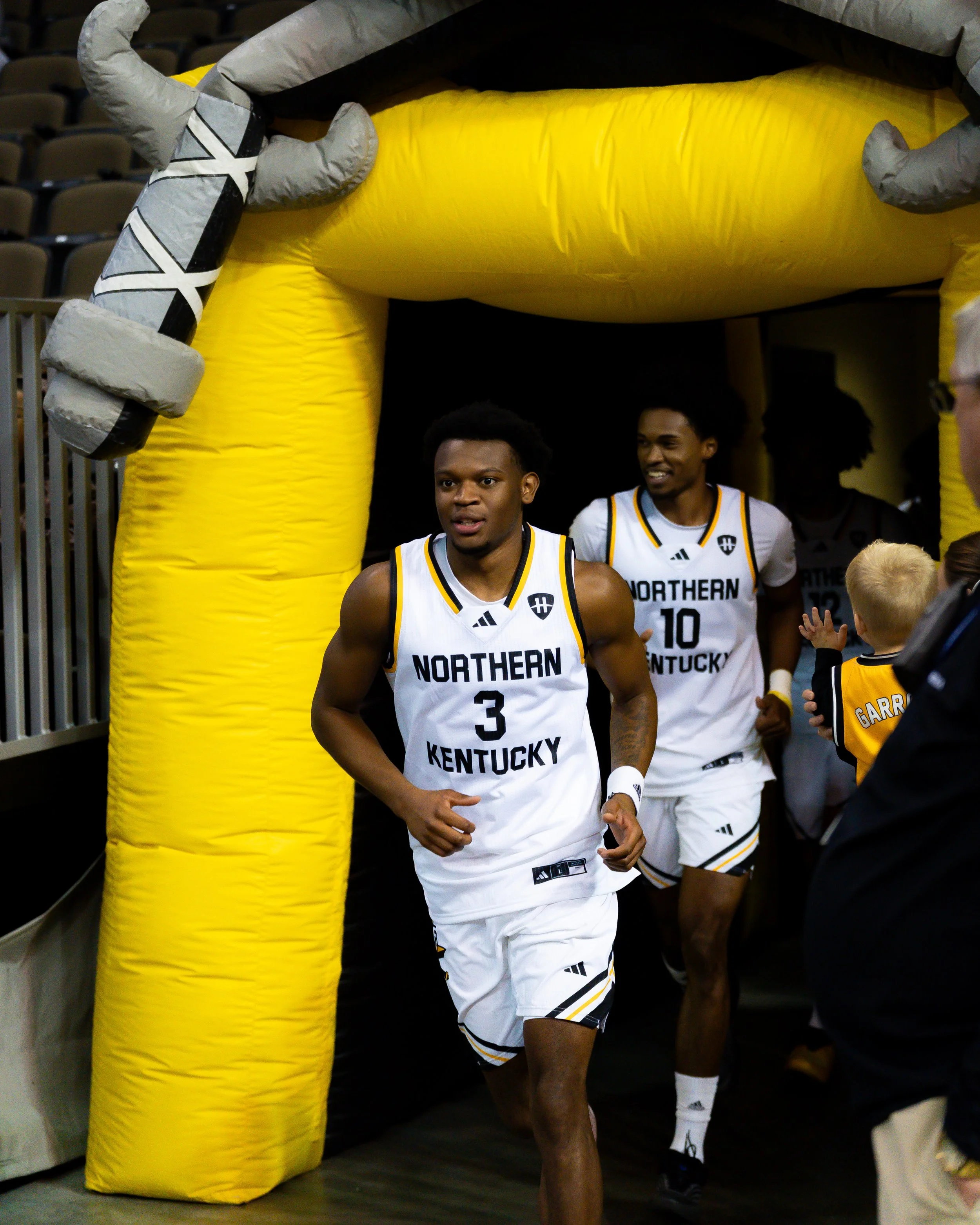 Northern Kentucky basketball players running through a tunnel onto the court in their white uniforms with black and yellow accents.
