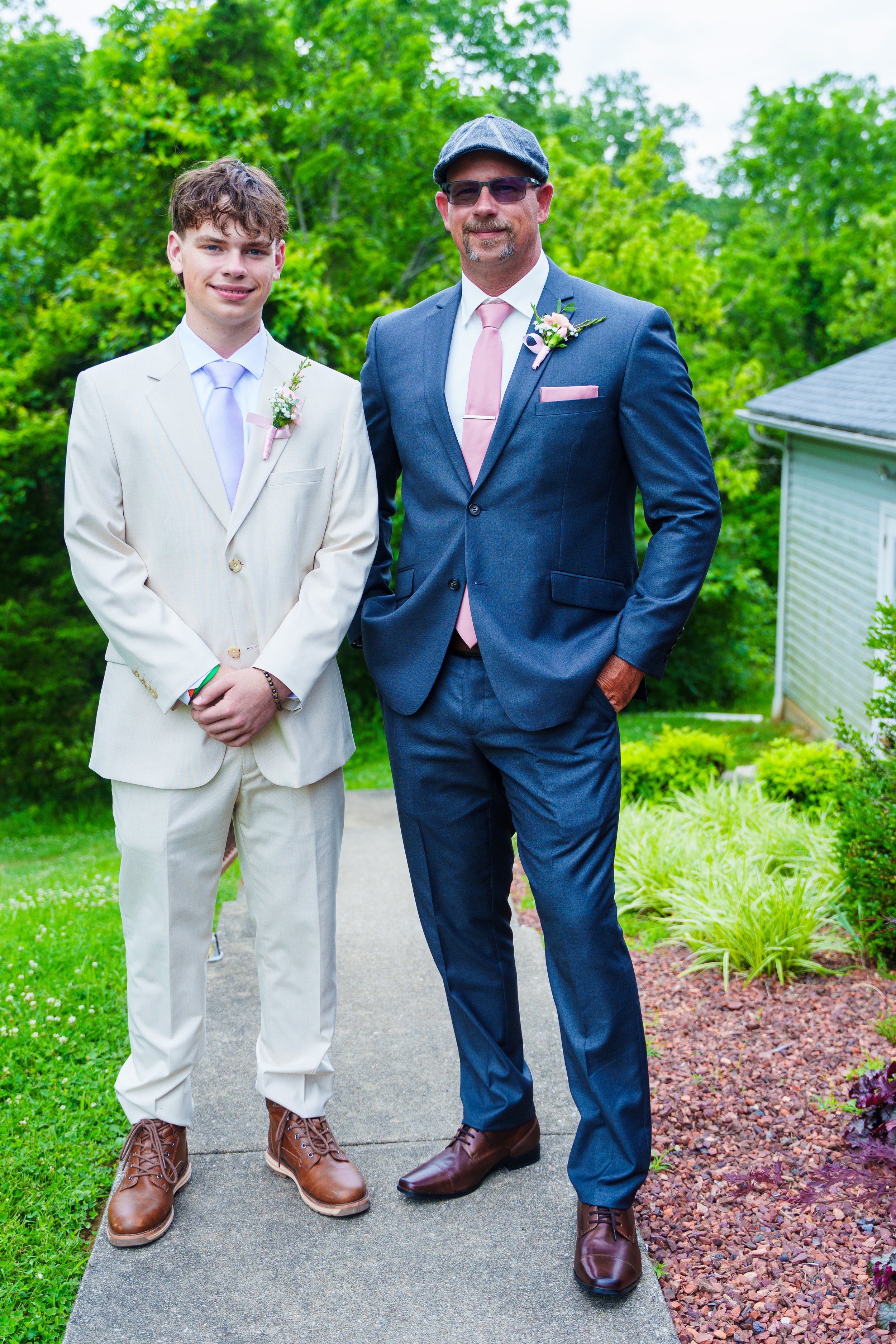 A young man and an older man stand outdoors on a sidewalk, dressed in formal suits with pink ties and boutonnières, surrounded by green foliage and flowering plants.