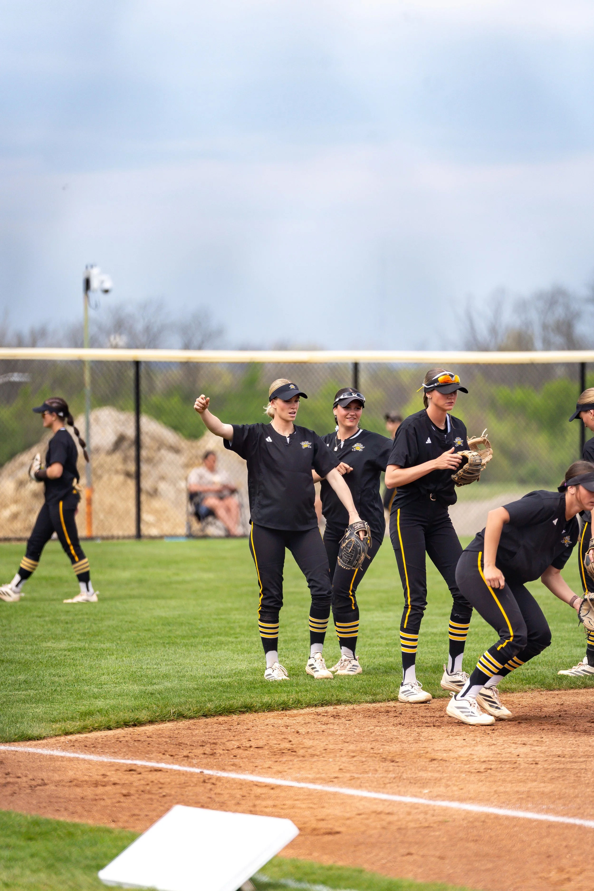 A group of female softball players in black uniforms with yellow stripe accents, practicing on the field. Some are standing, and others are kneeling or bending down near the baseline. One player is gesturing with her hand. They are wearing caps, sung