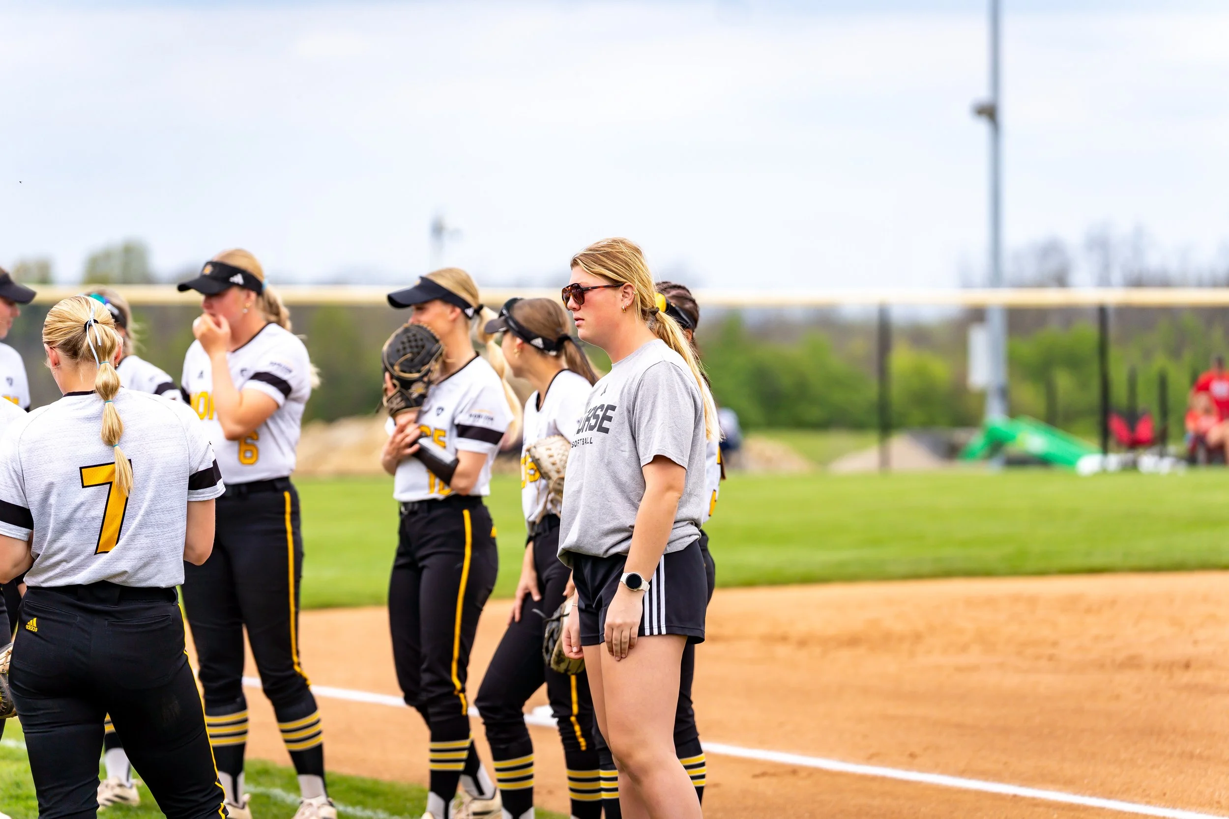 A group of female softball players standing on the field during a game or practice with some wearing white jerseys with yellow and black accents, and a coach or team member in a gray shirt and black shorts, all listening attentively.