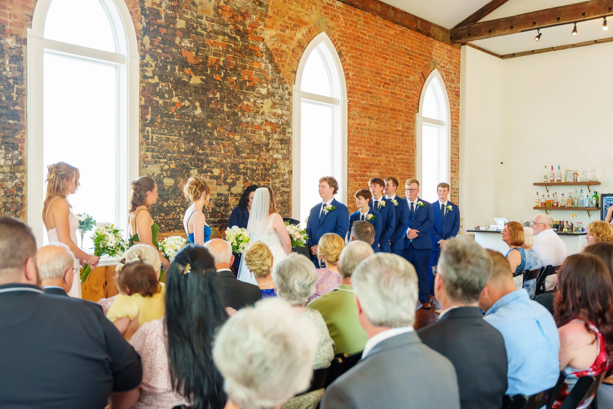A wedding ceremony with a bride and groom at the front of a room filled with guests. The room has exposed brick walls, large arched windows, and attendees sitting and watching the couple.