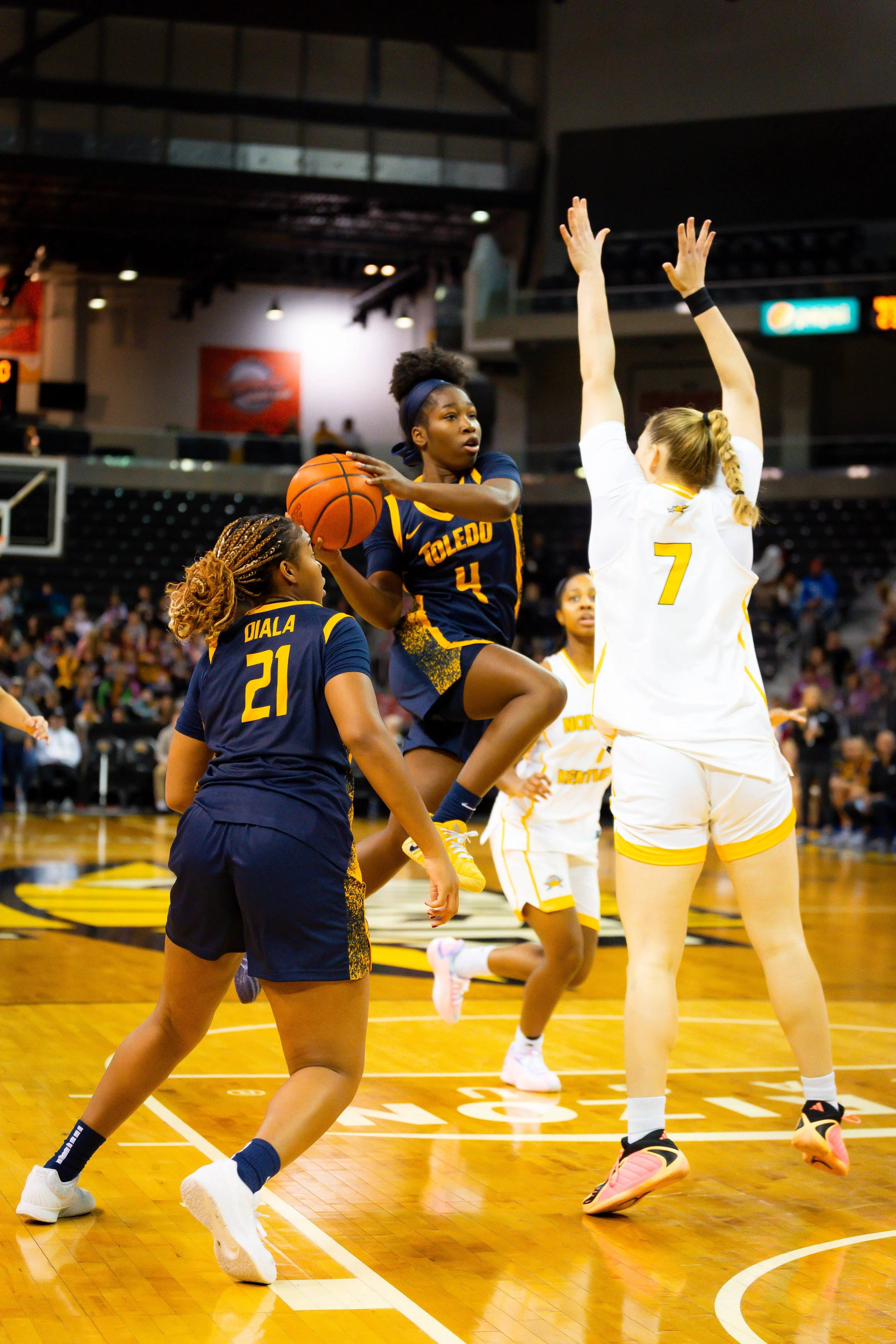 Women playing basketball in an indoor arena, with one player in a blue and yellow uniform jumping to shoot or pass, while an opponent in a white uniform with yellow accents attempts to block.