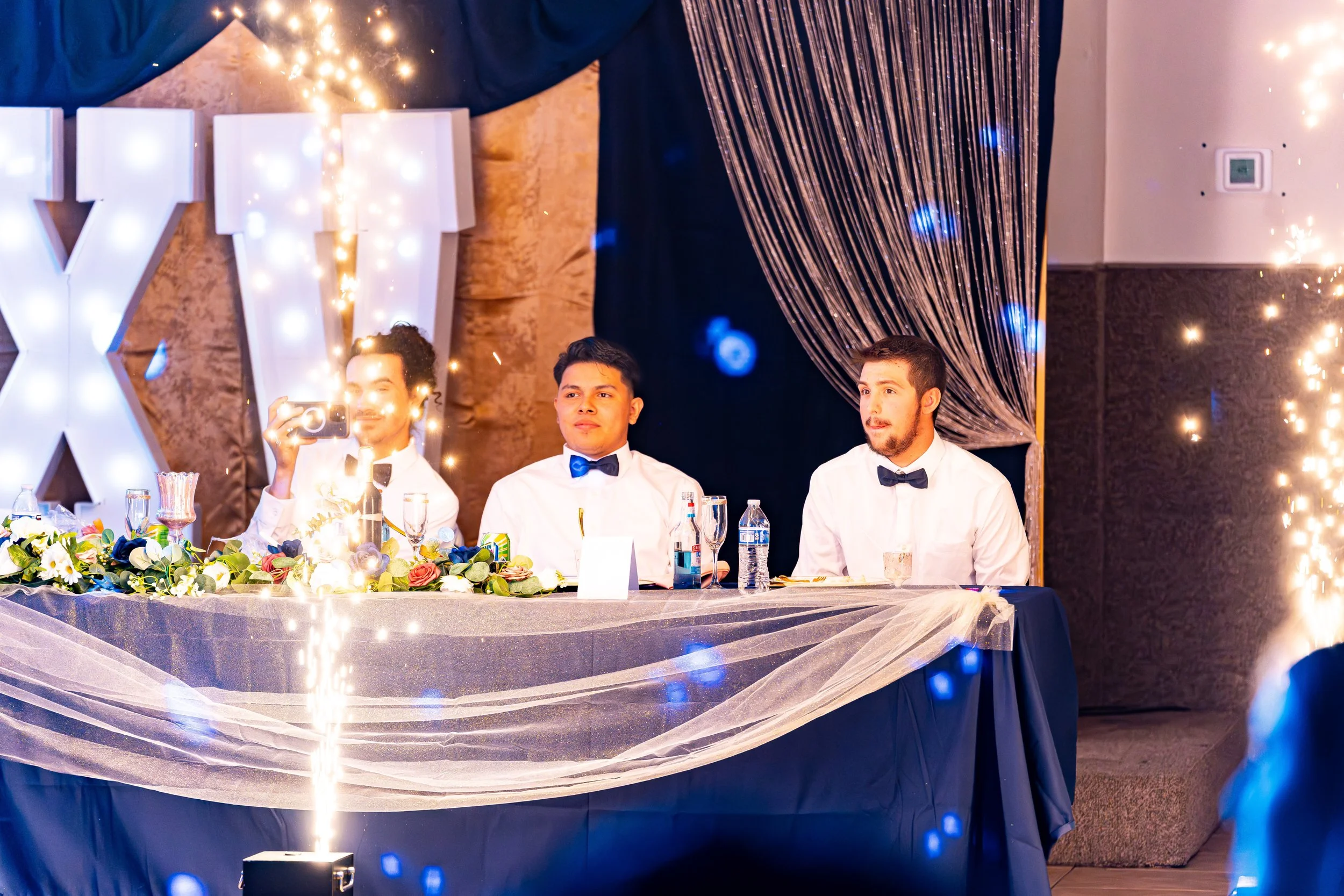 Three men dressed in formal attire sit at a decorated banquet table with flowers, water bottles, and glasses, during a celebration or wedding. A large illuminated letter formation and sparklers are visible in the background, with decorative drapes an